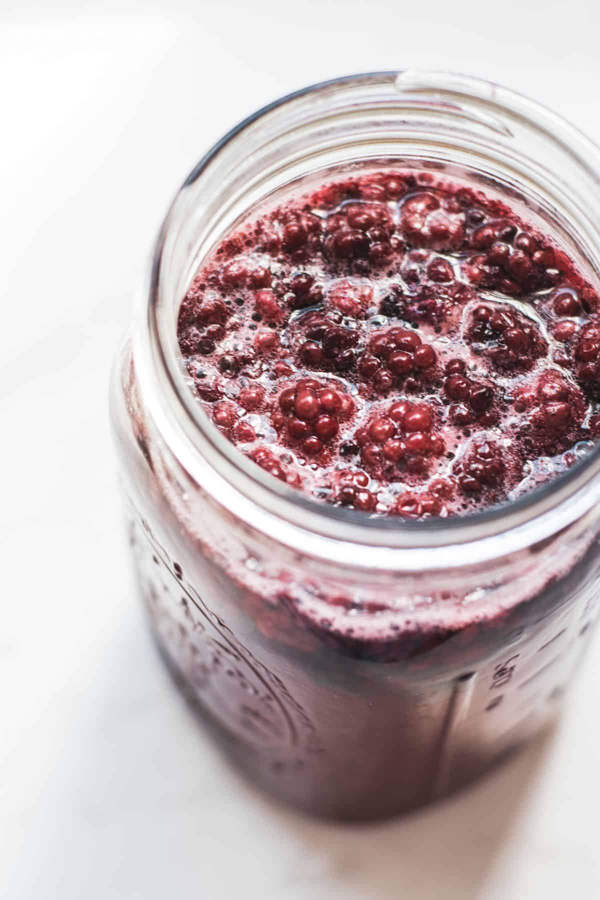A close-up of a glass jar filled with dark purple berries and juice, with bubbles and foam on the surface, sits on a white background—perfect for inspiring your next apple cider vinegar recipe.