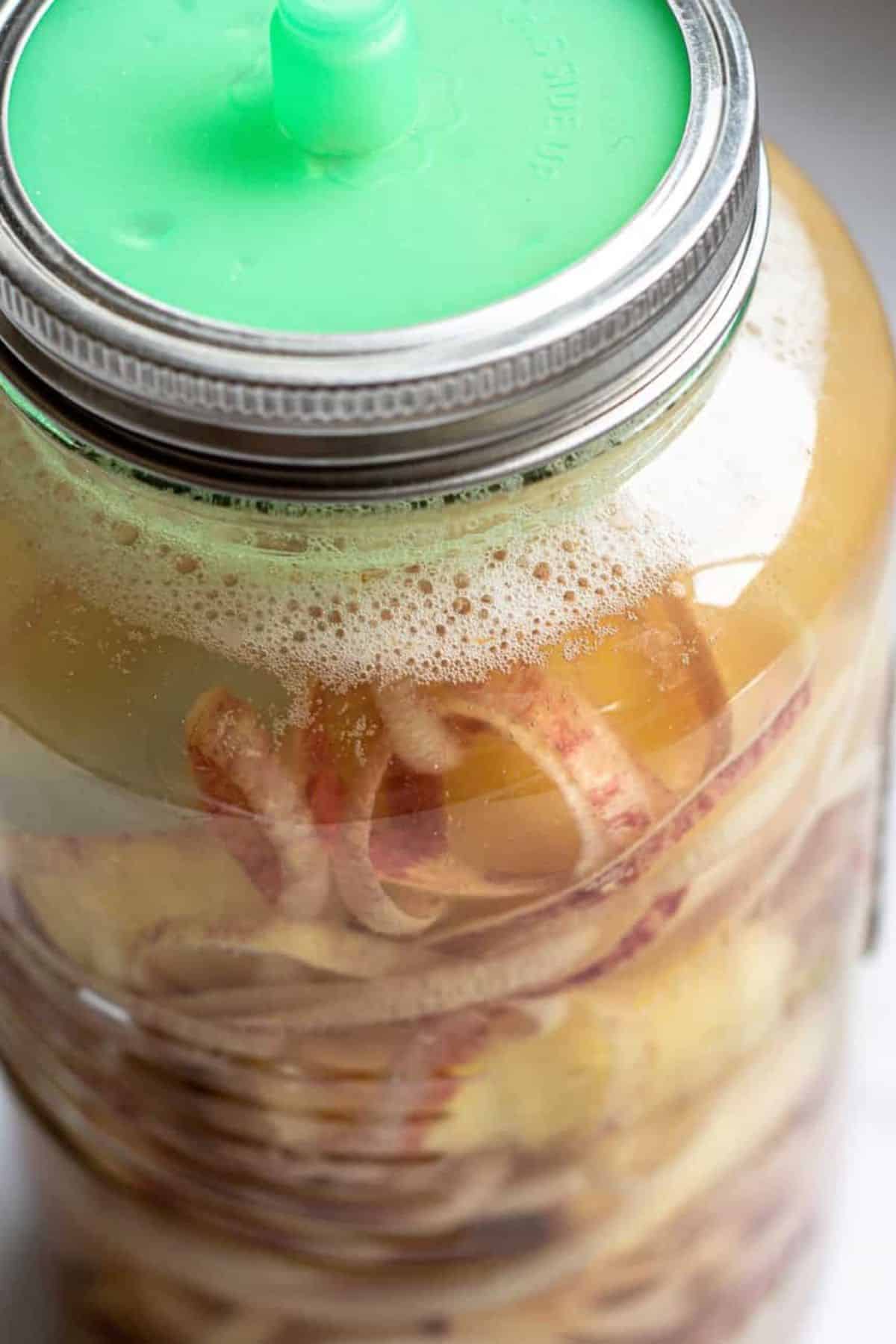 A close-up of a glass jar with a green fermenting lid, containing apple peels submerged in liquid with bubbles on the surface—perfect for starting your own apple cider vinegar recipe.