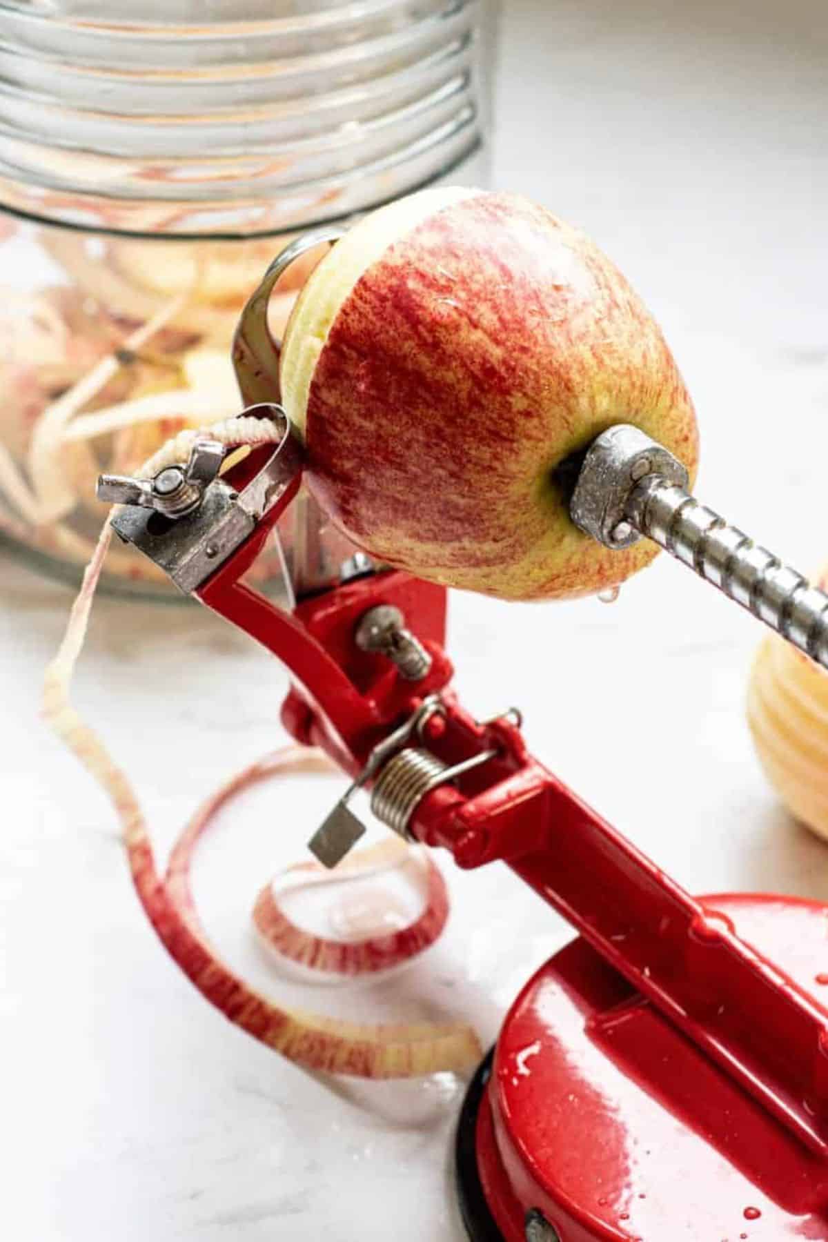 A red apple peeler and corer machine is peeling an apple, with the peel spiraling off. In the background, a glass jar filled with apple peels sits on a white surface—perfect for starting your next apple cider vinegar recipe.