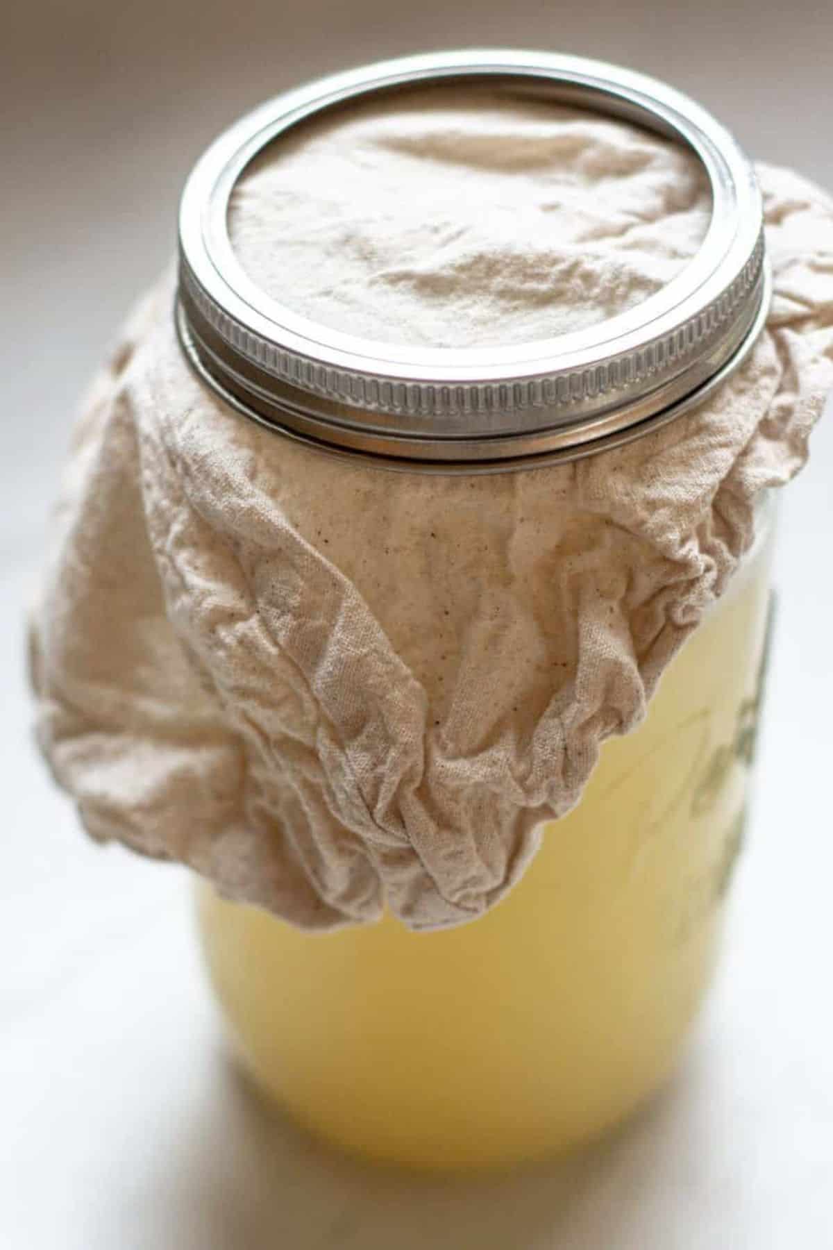 A glass mason jar filled with a pale yellow liquid, likely homemade from an apple cider vinegar recipe, is covered with a cloth secured by a metal ring lid, perfect for fermentation or straining.