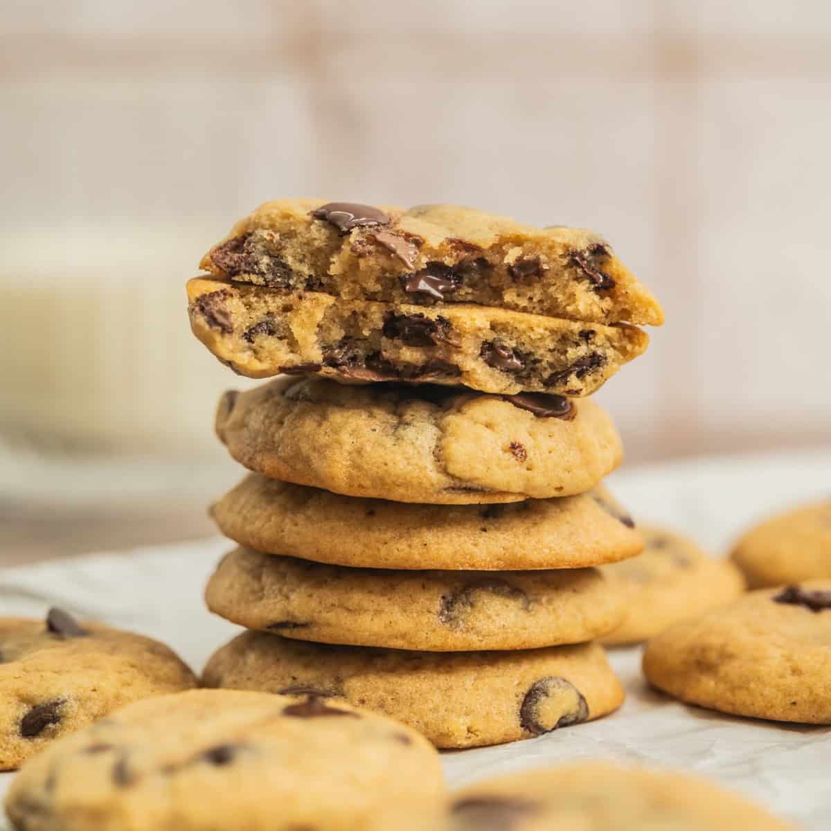A stack of Sourdough Chocolate Chip Cookies sits on parchment paper, with the top cookie broken in half to reveal a gooey chocolate center. More cookies are scattered in the background.