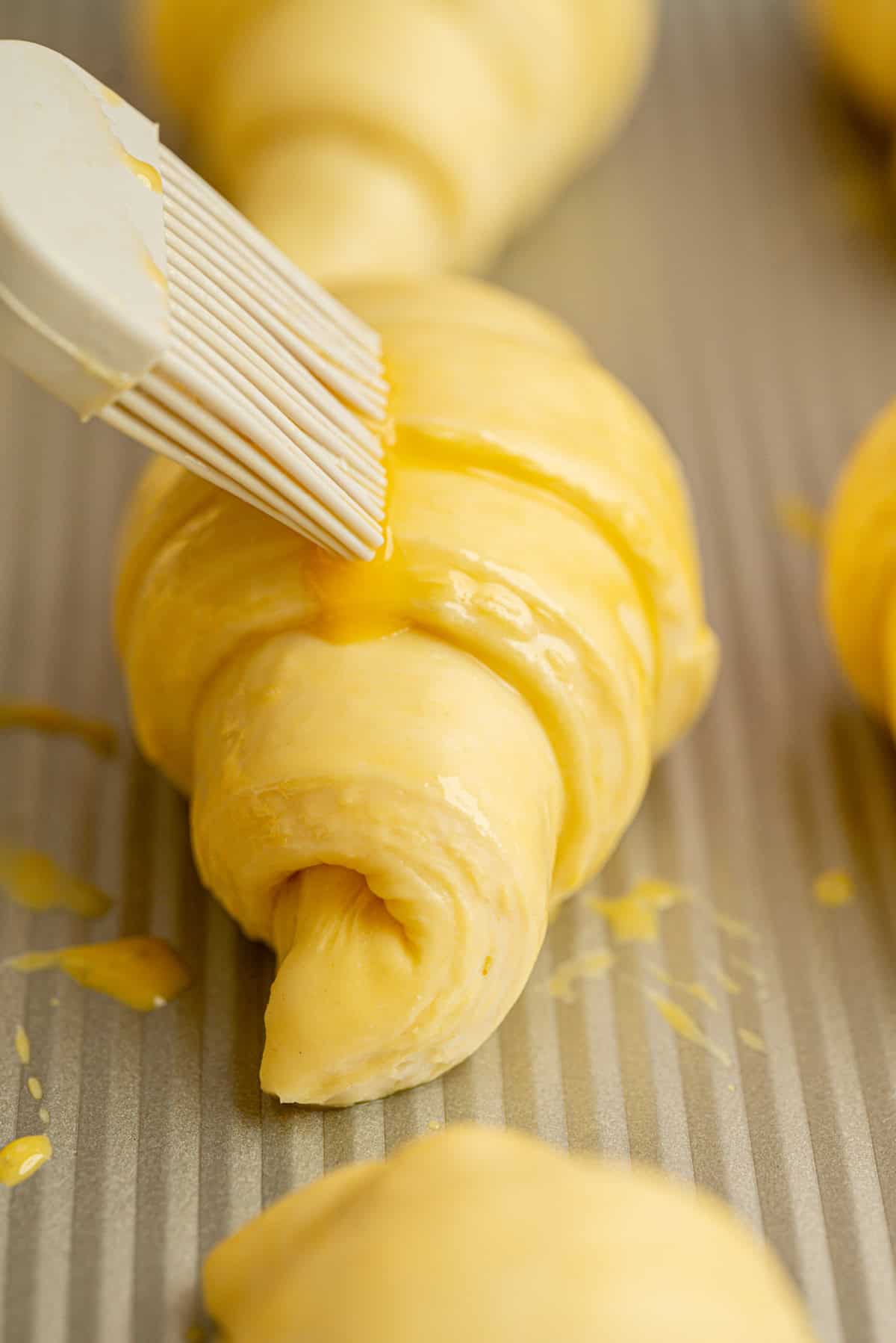 A pastry brush applies an egg wash to unbaked sourdough croissants on a baking sheet, giving the pastries a shiny, golden coating.