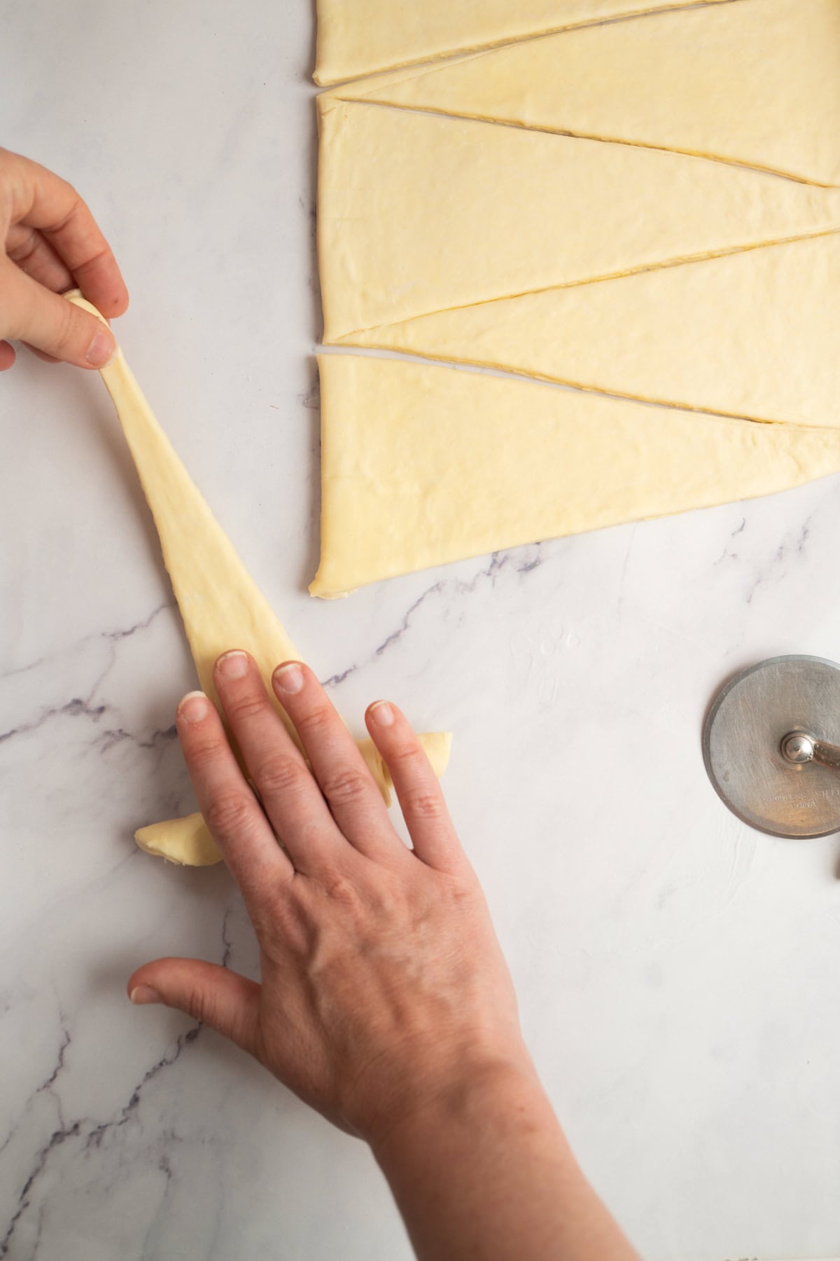 A person stretching and rolling a triangular piece of dough for sourdough croissants on a marble surface, with a pastry cutter and other dough triangles nearby.