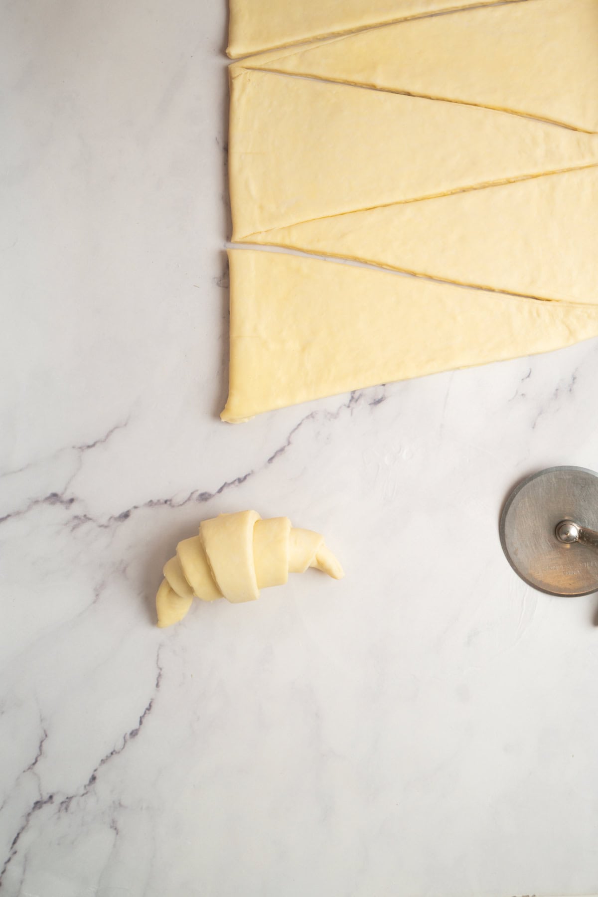 A sheet of rolled-out dough cut into triangles for sourdough croissants, a single unbaked croissant, and a pizza cutter rest on a marble countertop.