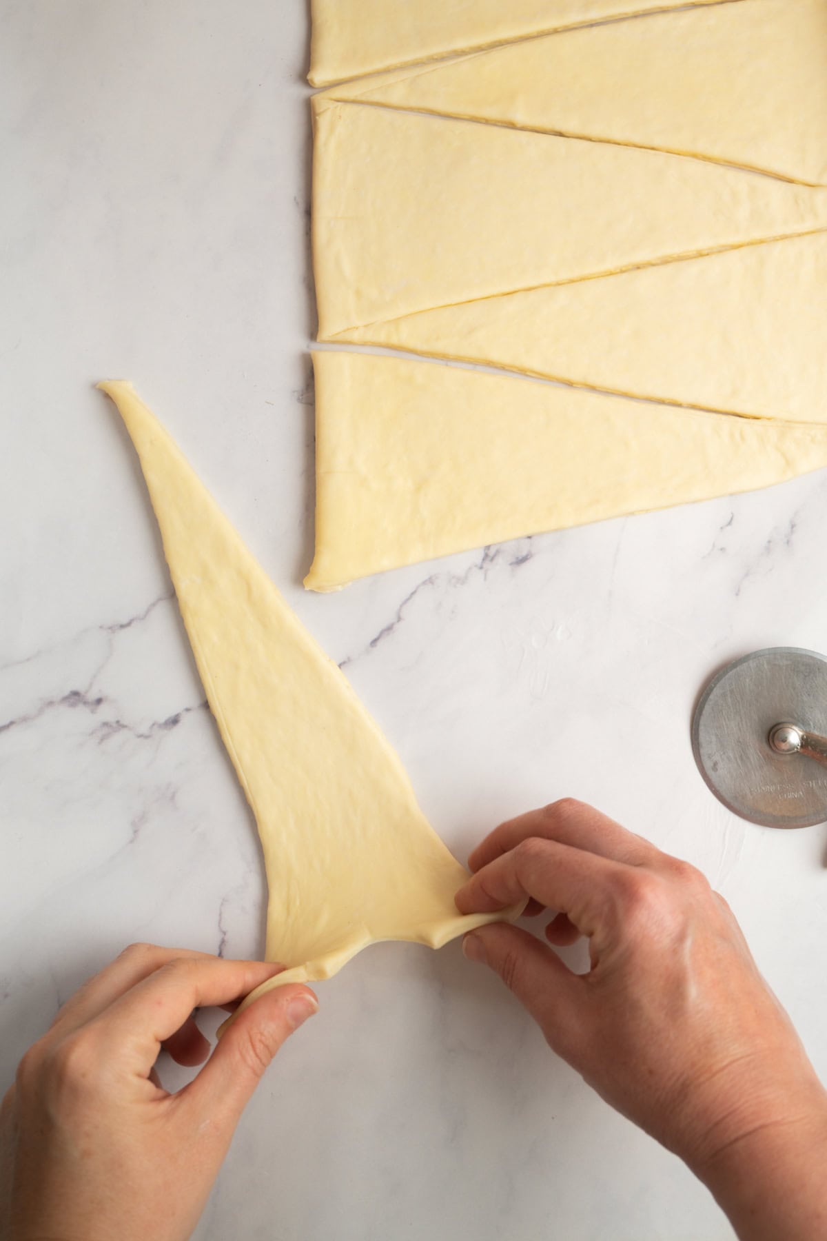 Hands stretching a triangle of dough cut from a larger sheet, preparing sourdough croissants, with more dough triangles and a pastry wheel on a marble surface.