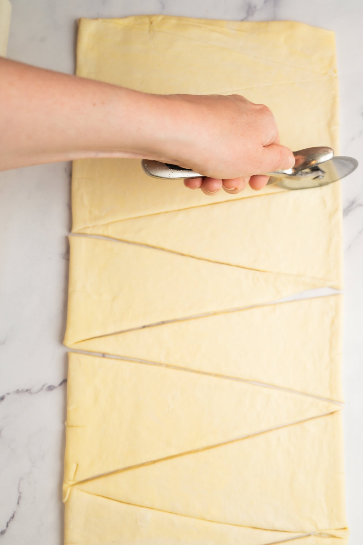 A hand uses a pizza cutter to slice a sheet of dough for sourdough croissants into long, triangular strips on a marble surface.