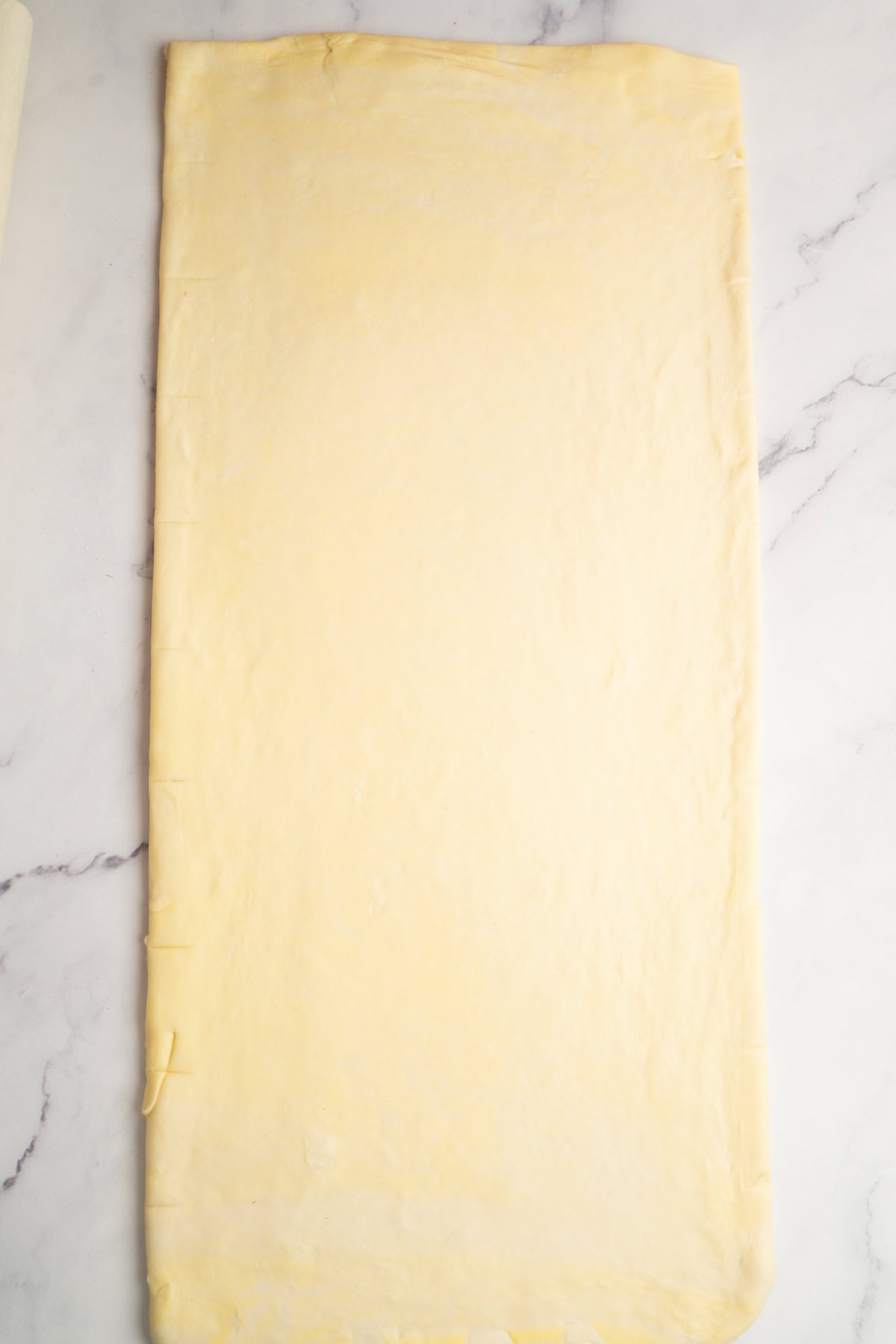 A large rectangular sheet of rolled-out puff pastry dough, perfect for making sourdough croissants, rests on a white marble surface.