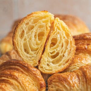 A golden, flaky sourdough croissants cut in half reveals its airy, layered interior, resting atop whole sourdough croissants on a cooling rack with a blurred background.