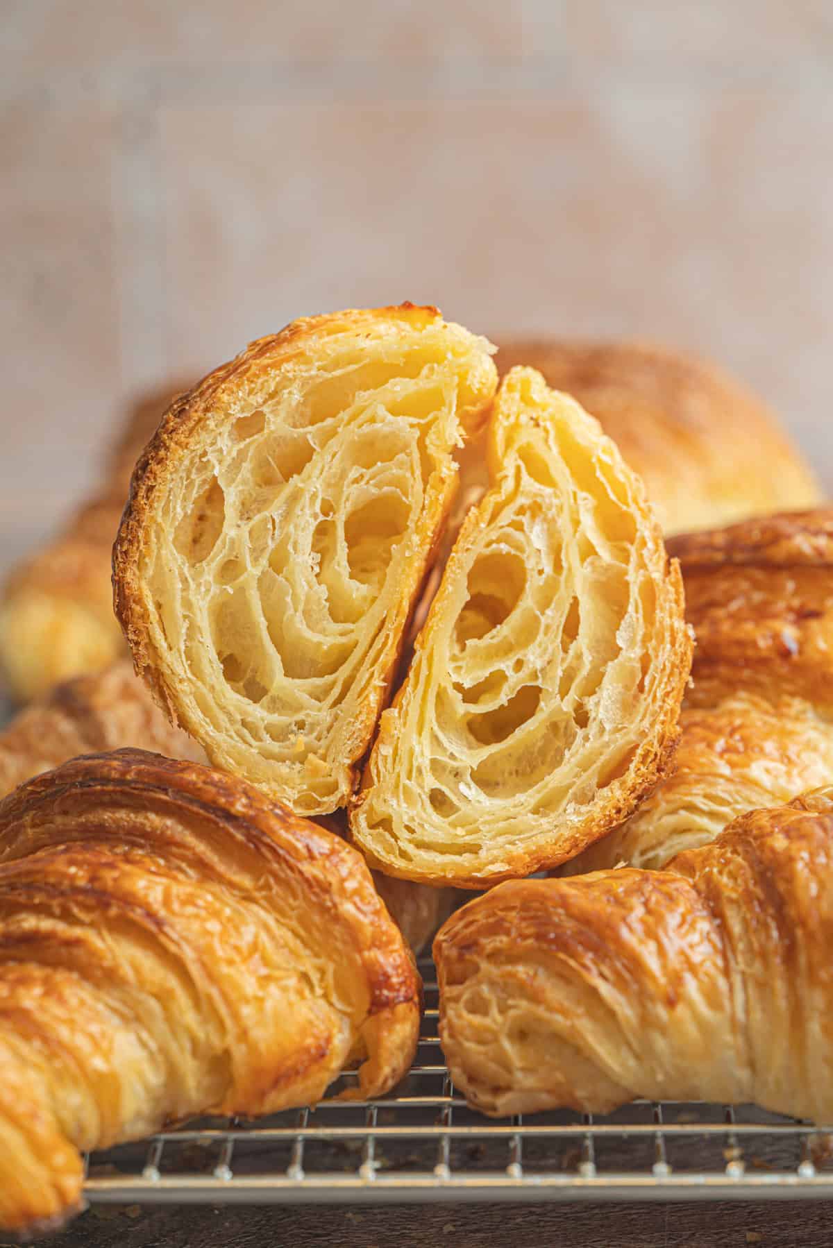 A golden, flaky sourdough croissant cut in half reveals its airy, layered interior, resting atop whole sourdough croissants on a cooling rack with a blurred background.