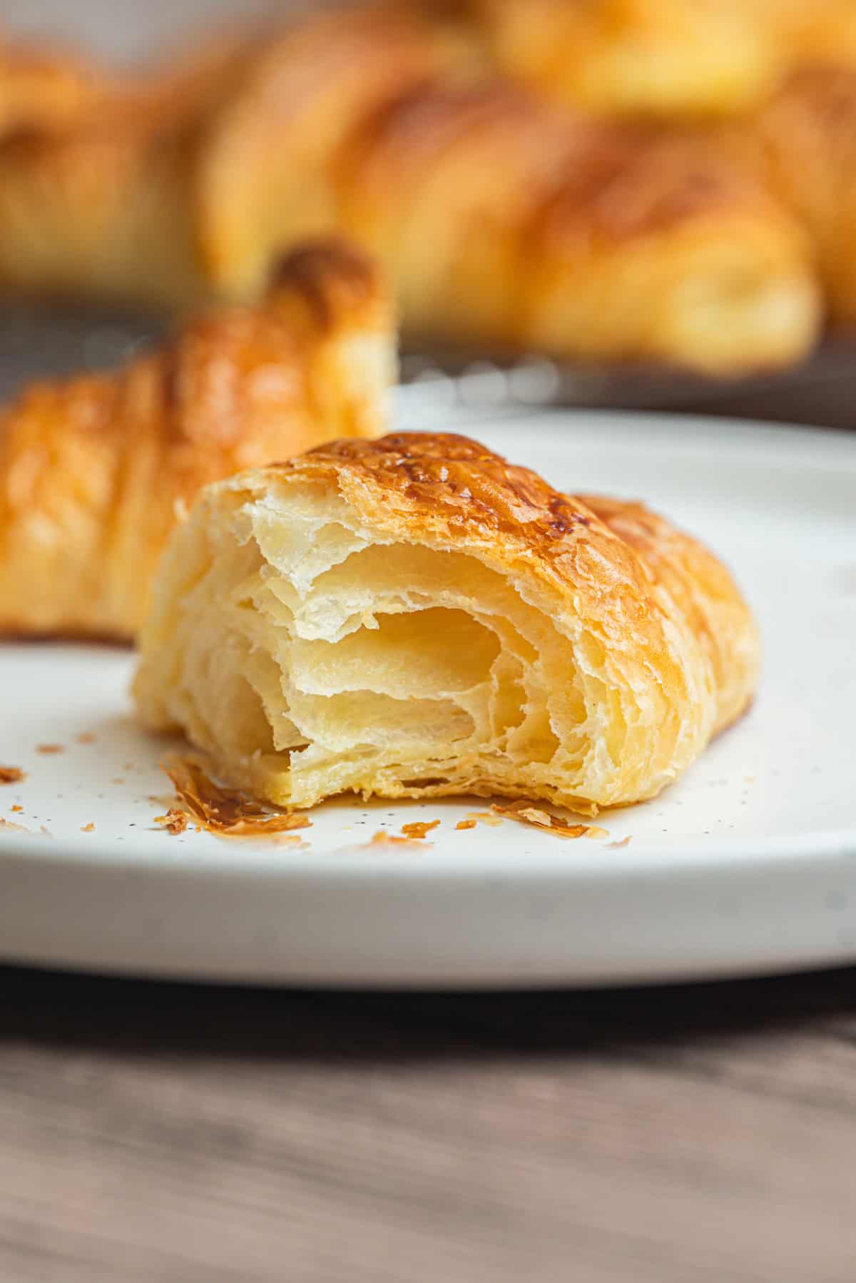 A close-up of a flaky, golden sourdough croissant with a bite taken out, showing its airy layers. The croissant sits on a white plate, with more pastries blurred in the background.