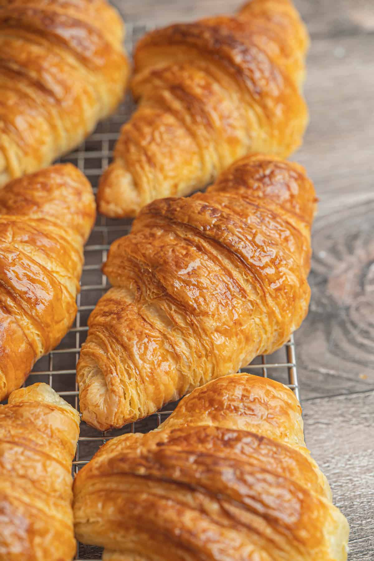 Golden, flaky sourdough croissants cooling on a metal wire rack, resting on a rustic wooden surface. The croissants have a shiny, crisp outer layer with visible, delicate layers.