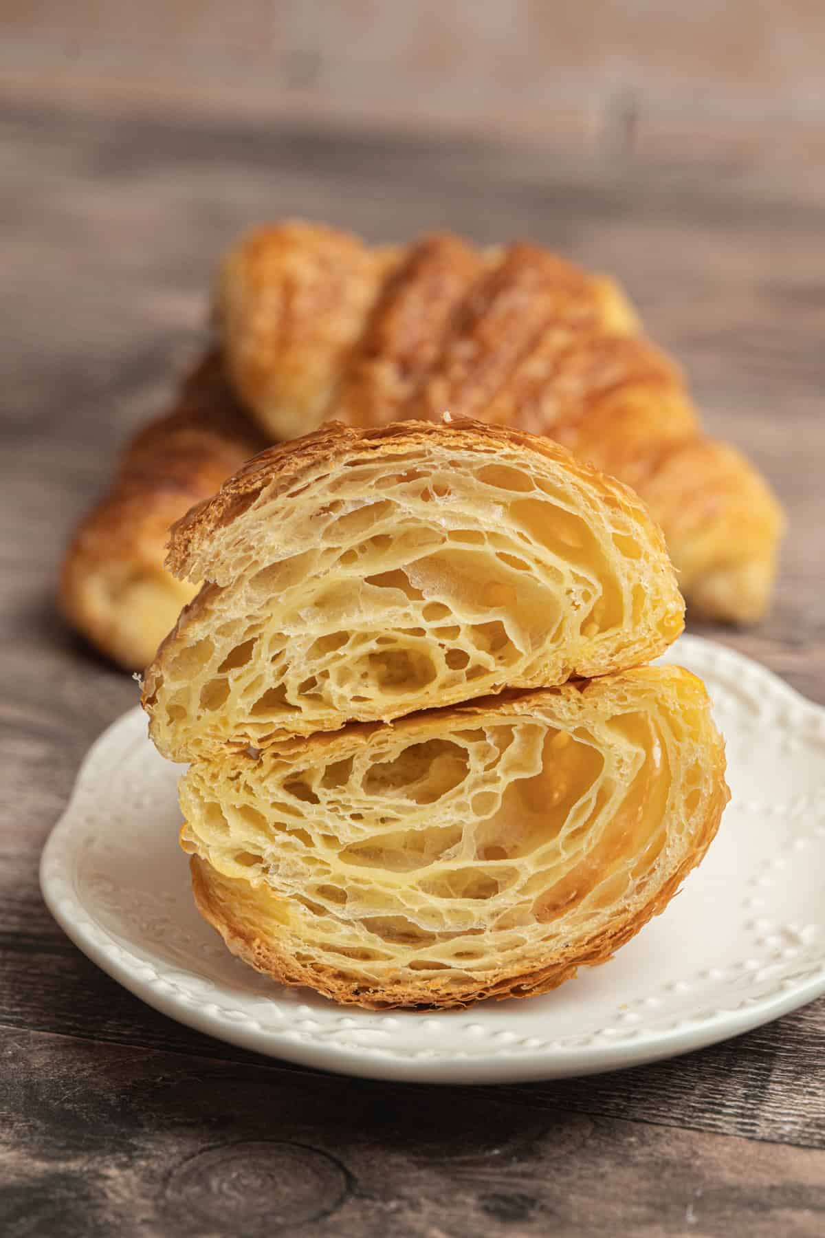 A flaky sourdough croissant cut in half, revealing its airy, layered interior, sits on a white plate with two whole croissants blurred in the background on a wooden surface.