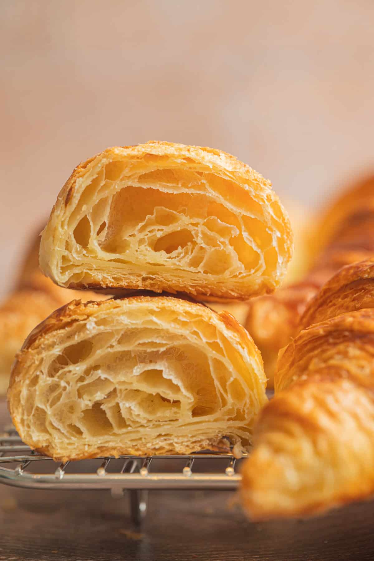 A close-up of a sourdough croissant cut in half, revealing its airy, flaky, and golden layers, resting on a cooling rack with more sourdough croissants blurred in the background.