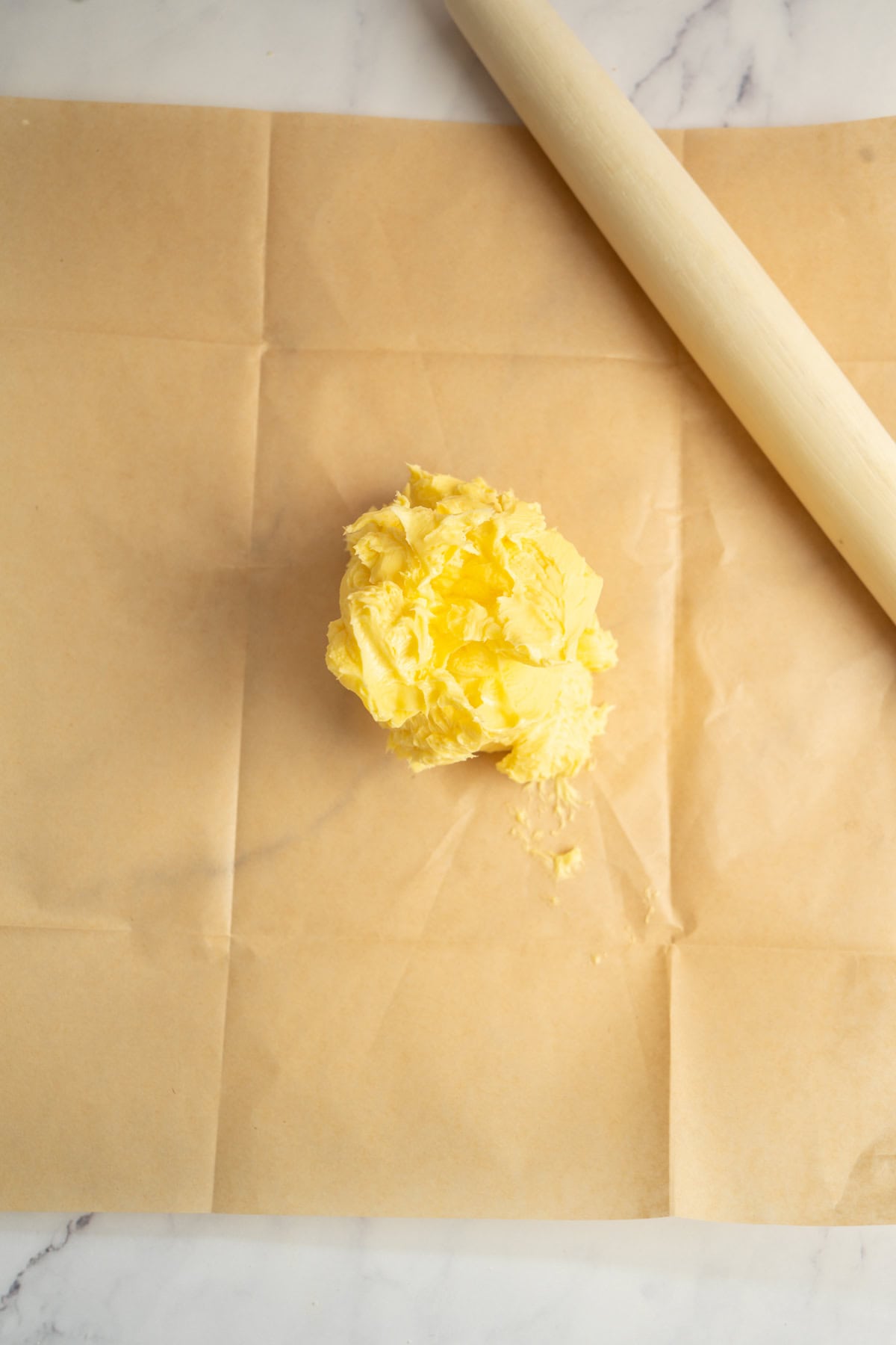 A mound of yellow butter sits on a sheet of brown parchment paper next to a light-colored rolling pin—ready for baking fresh sourdough croissants—on a marble surface.