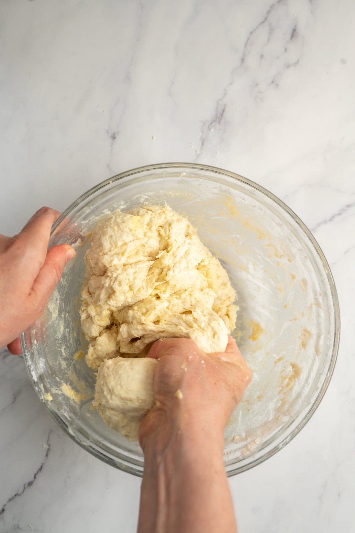 Hands kneading dough for sourdough croissants in a glass bowl on a white marble surface.