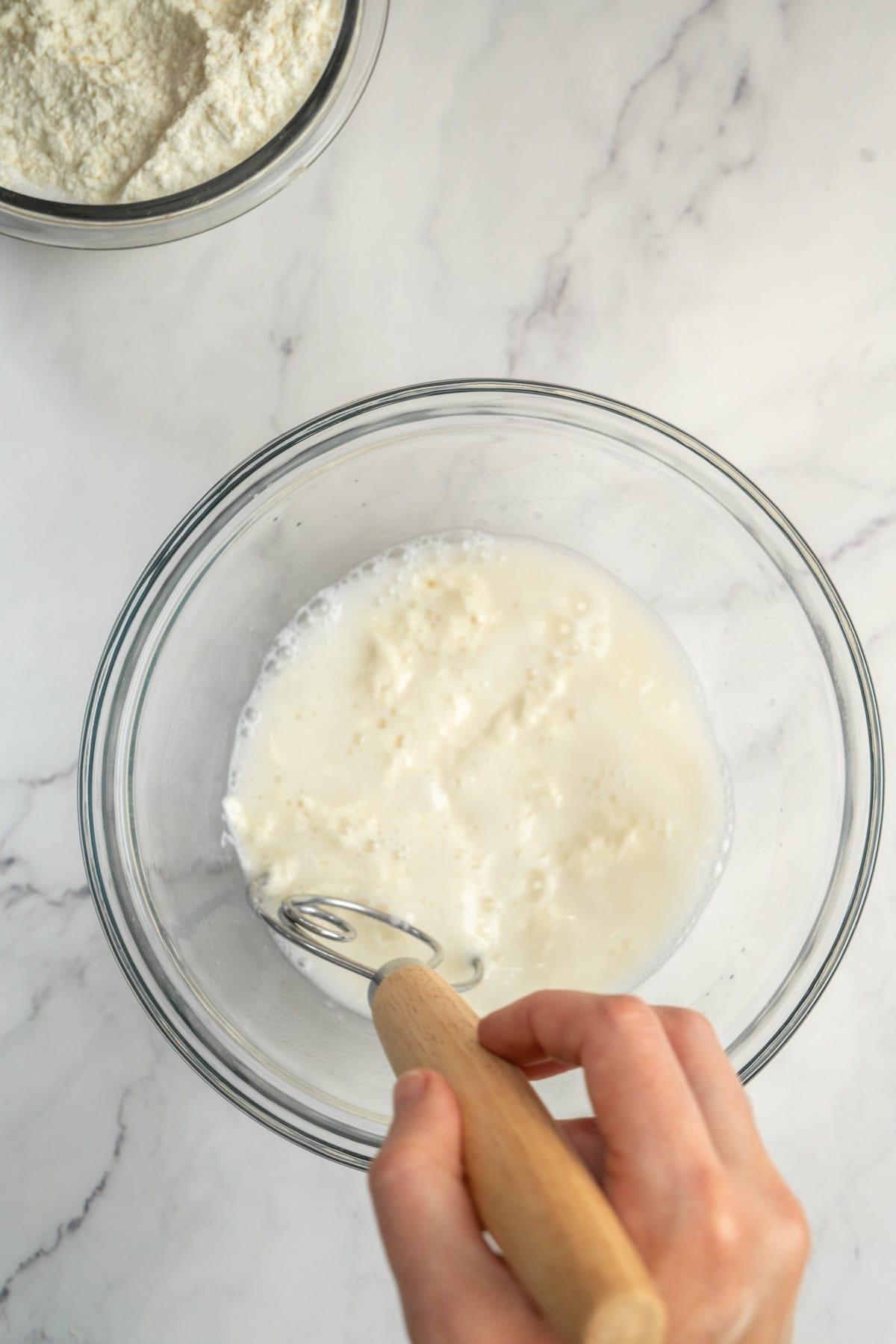 A hand uses a whisk to mix a white liquid in a clear glass bowl on a marble countertop—a key step when preparing sourdough croissants. Another bowl with white powder, possibly flour, sits in the top left corner.