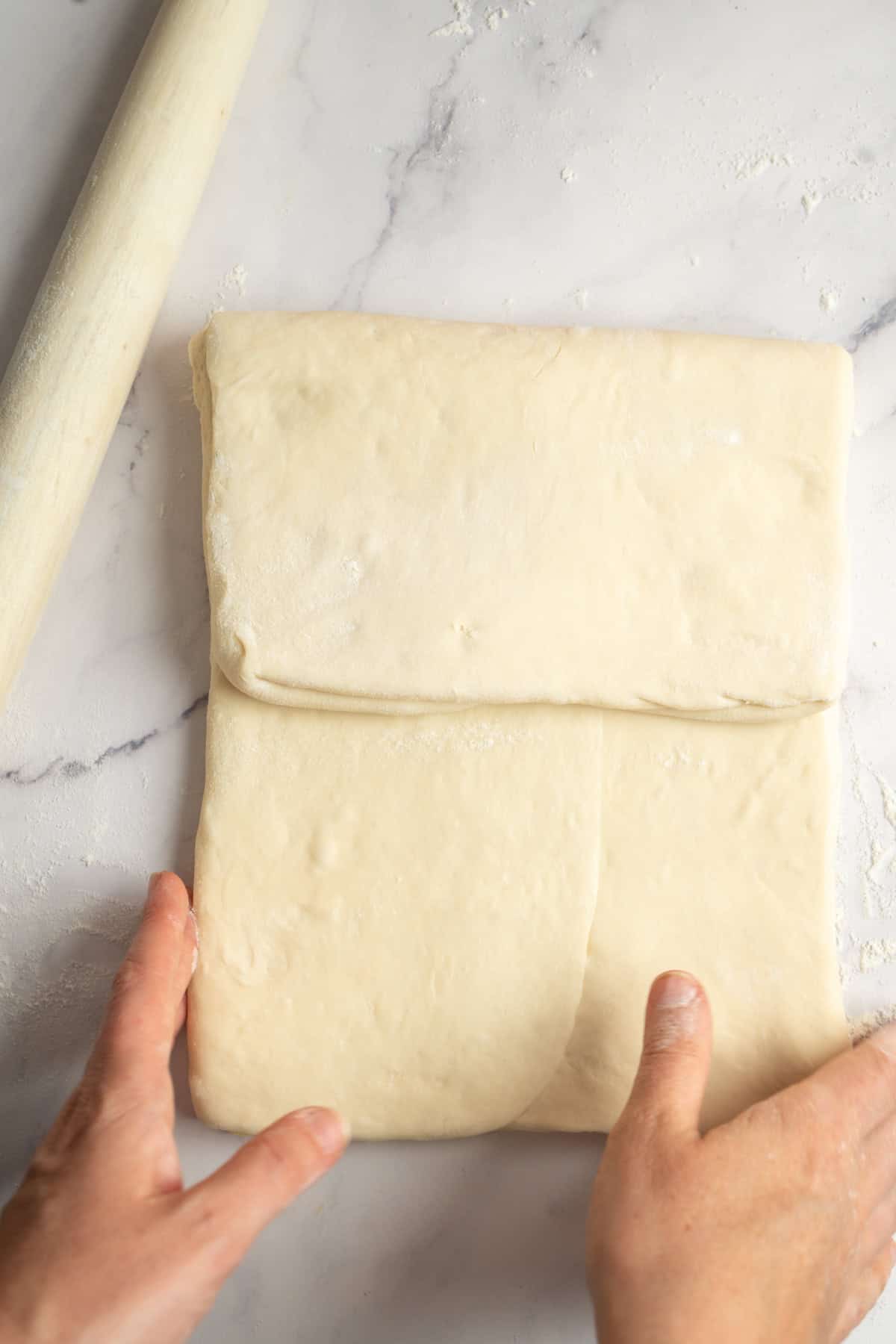 Hands folding a sheet of dough for sourdough croissants on a floured marble surface, with a rolling pin visible nearby.