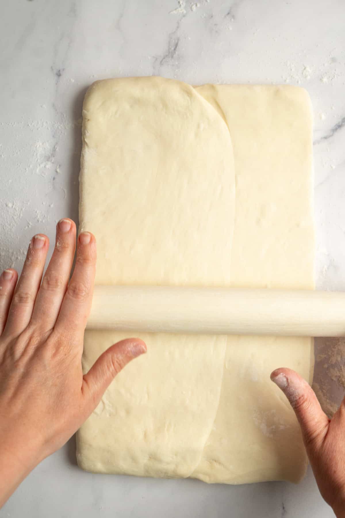 Hands using a rolling pin to roll out folded sourdough croissant dough on a floured, light-colored surface.