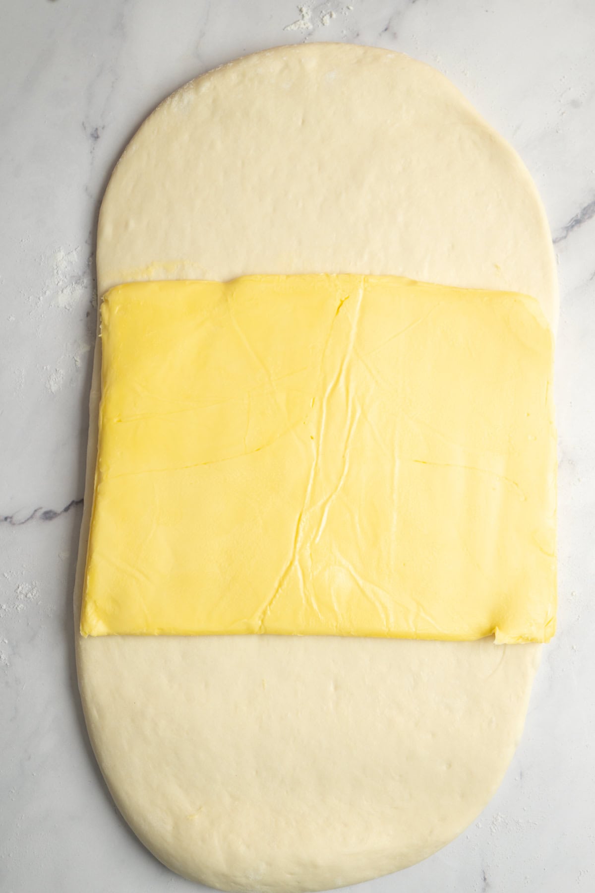 A rectangular sheet of butter is placed in the center of a large oval of rolled-out dough on a white marble surface, ready to be transformed into flaky sourdough croissants.