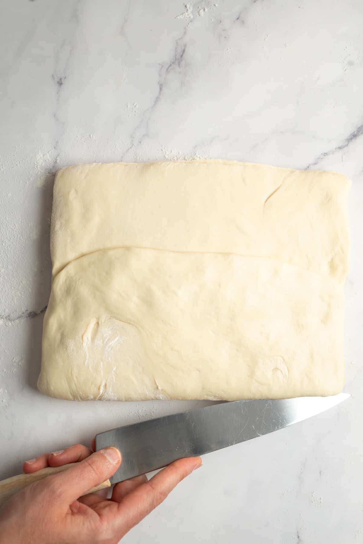 A hand holding a knife is about to cut a folded sheet of dough for sourdough croissants on a white marble surface.
