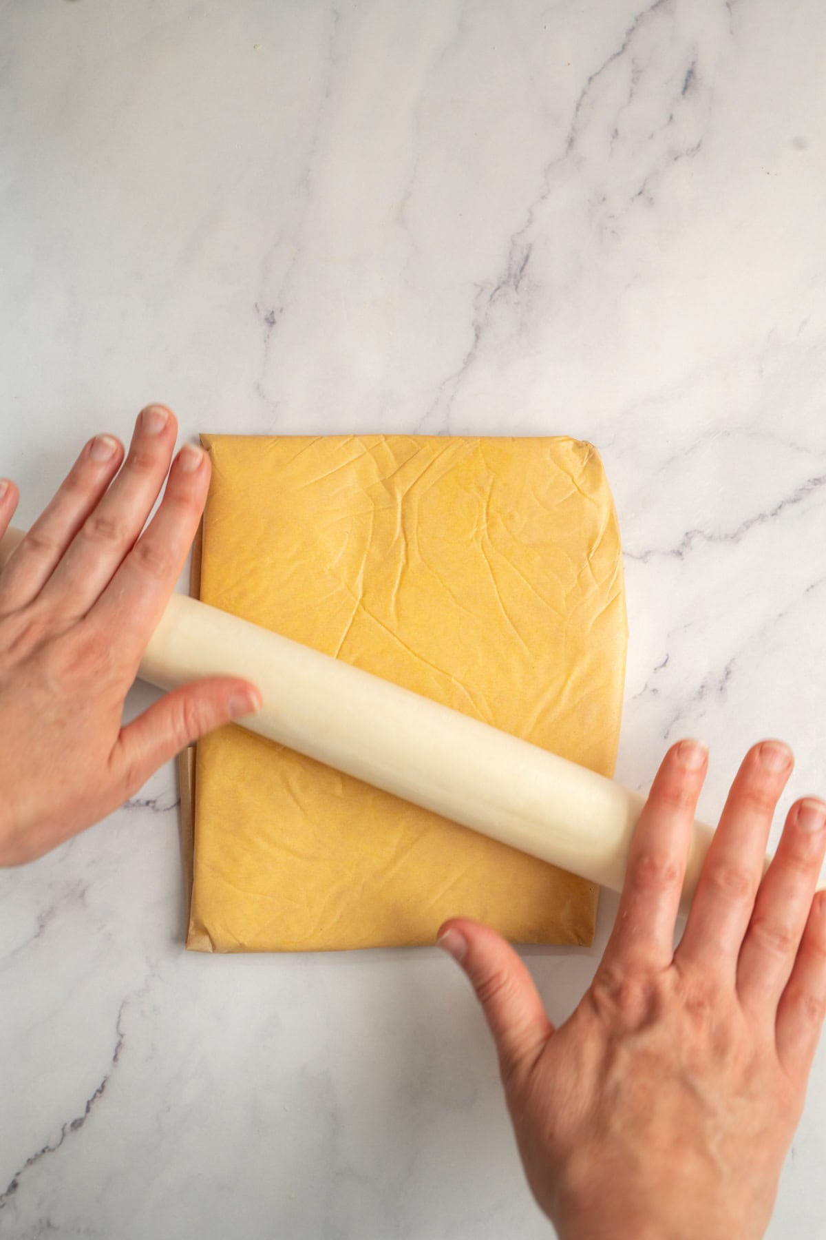 Two hands use a white rolling pin to flatten a square of yellow sourdough croissant dough on a light marble surface.