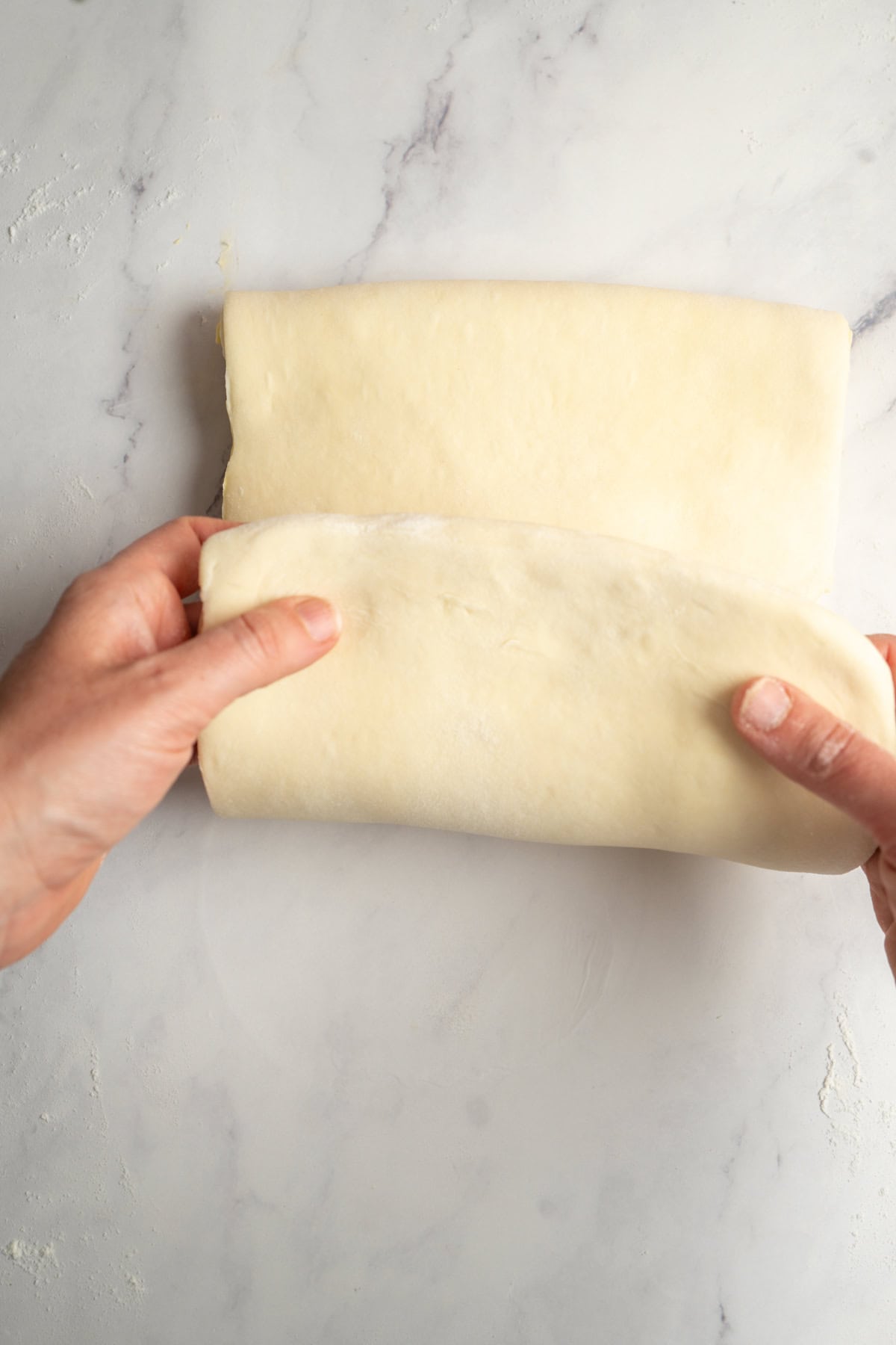 Two hands folding a sheet of pale dough on a lightly floured, white marble surface, preparing delicate sourdough croissants for baking.