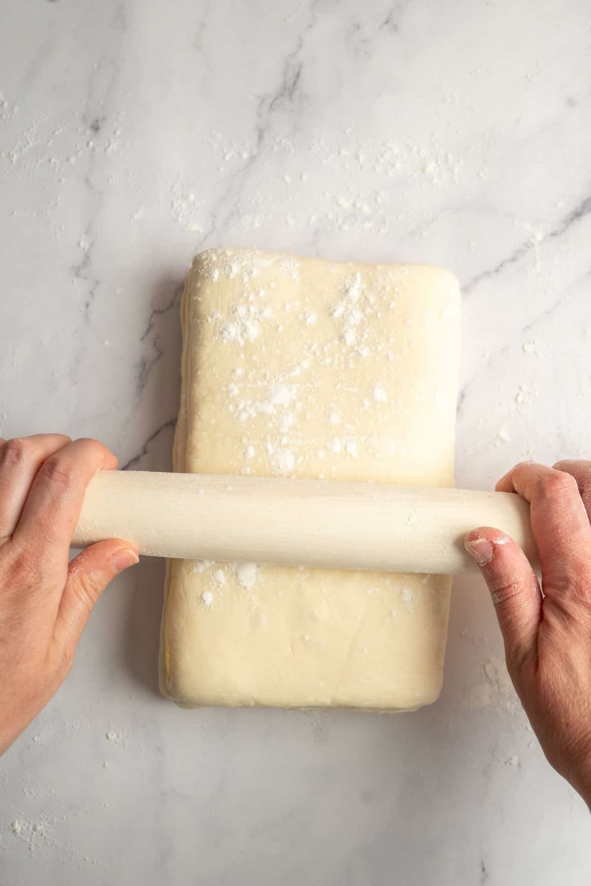 Hands rolling out a rectangular block of dough with a rolling pin on a floured, light-colored surface, preparing perfect sourdough croissants.