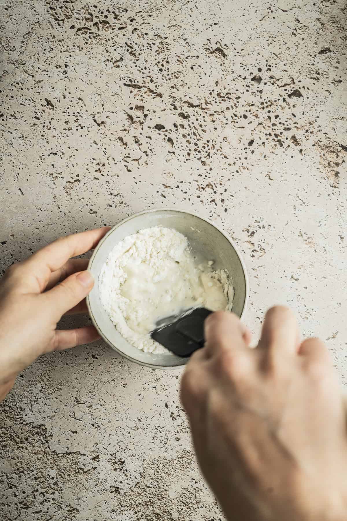 A person stirs flour and liquid together in a small bowl with a black spatula on a textured light-colored surface, demonstrating how to make sourdough starter.