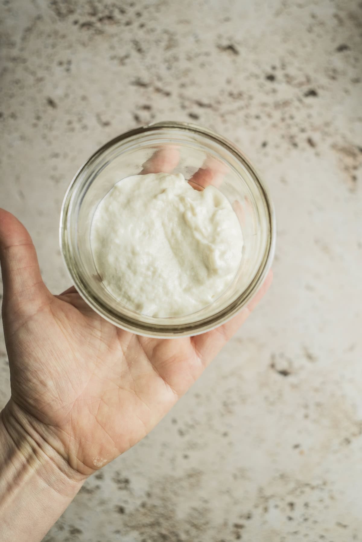 A hand holds a small glass bowl containing a creamy white substance, possibly yogurt or whipped cream, against a light, speckled background—perfect for those curious about how to make sourdough starter from scratch.
