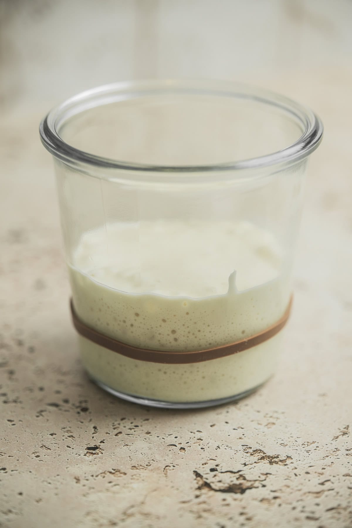 A clear glass jar partially filled with creamy, bubbly sourdough starter sits on a light, textured surface—an essential step in learning how to make sourdough starter. A brown rubber band is wrapped around the jar.