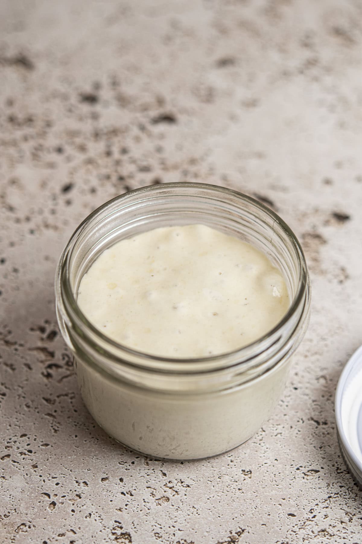 An open glass jar filled with sourdough starter sits on a speckled countertop, reminiscent of jars often used for how to make sourdough starter. The white lid rests nearby on the right side.