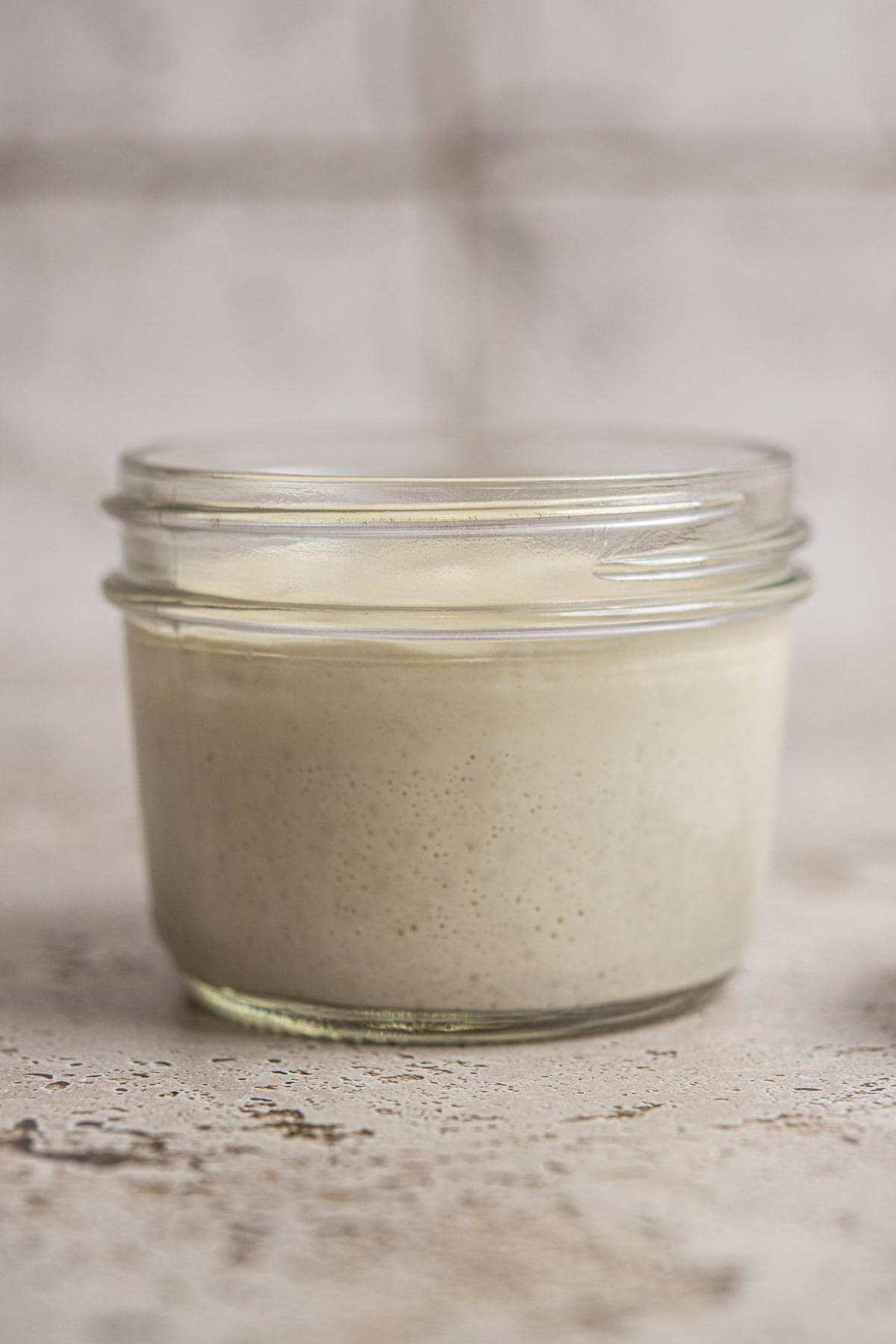 A small glass jar filled with creamy, light beige sourdough starter—a perfect visual for anyone curious about how to make sourdough starter—rests on a textured surface, the background softly blurred.