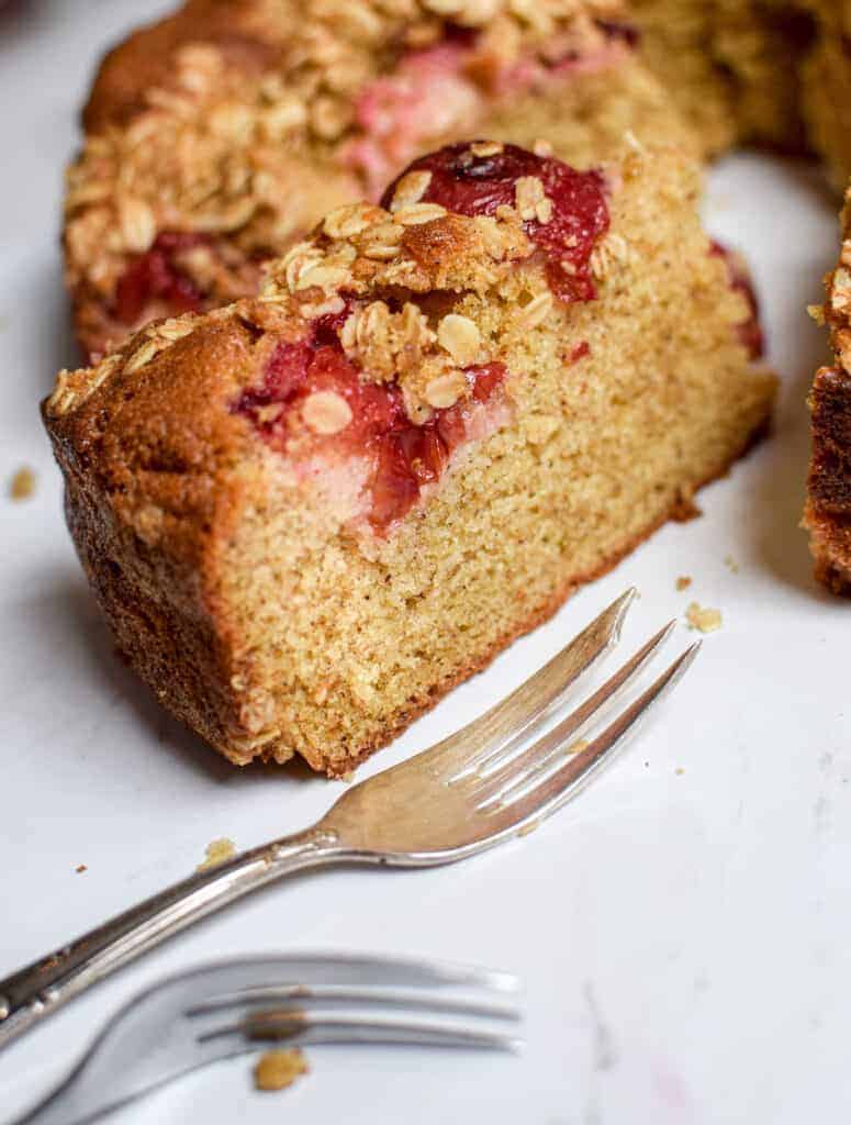 a close up of a cake slice with oats on top and red Plum showing. Two cake forks are in the foreground 