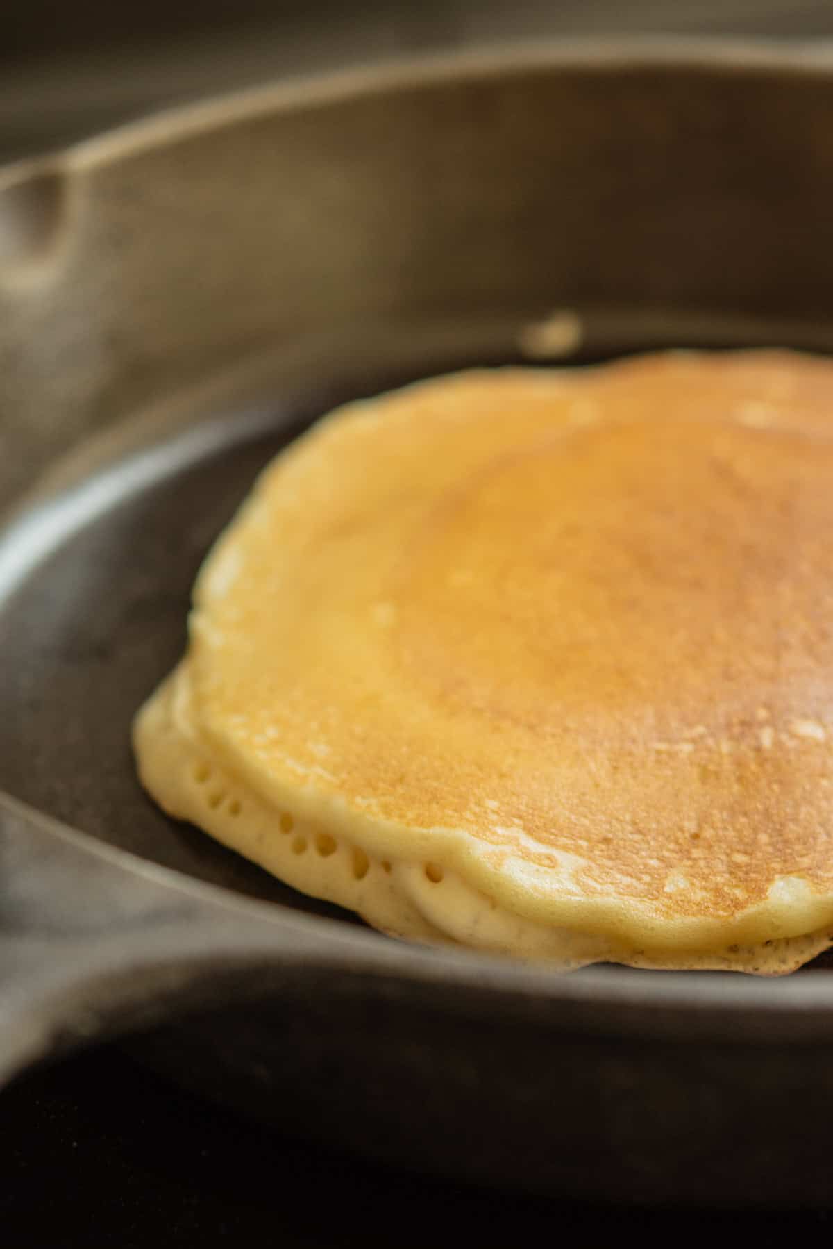 A golden-brown sourdough discard pancake cooks in a black cast iron skillet, its edges slightly lifting and bubbles forming along the side—clear signs this tangy treat is nearly ready to be flipped.