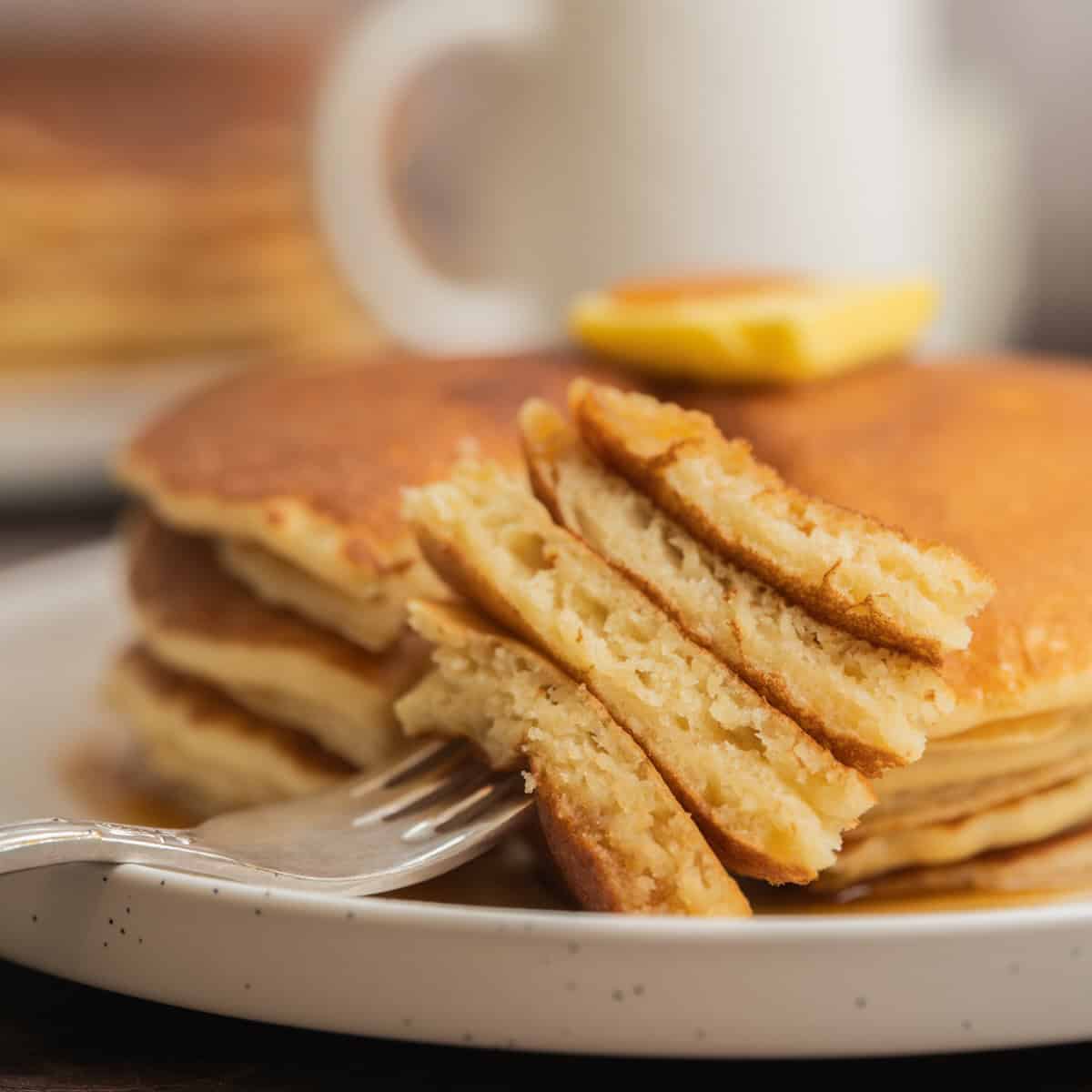 A close-up of a stack of fluffy sourdough discard pancakes on a white plate, with a pat of butter on top and a fork holding a bite-sized piece in the foreground. A mug and more pancakes are blurred in the background.