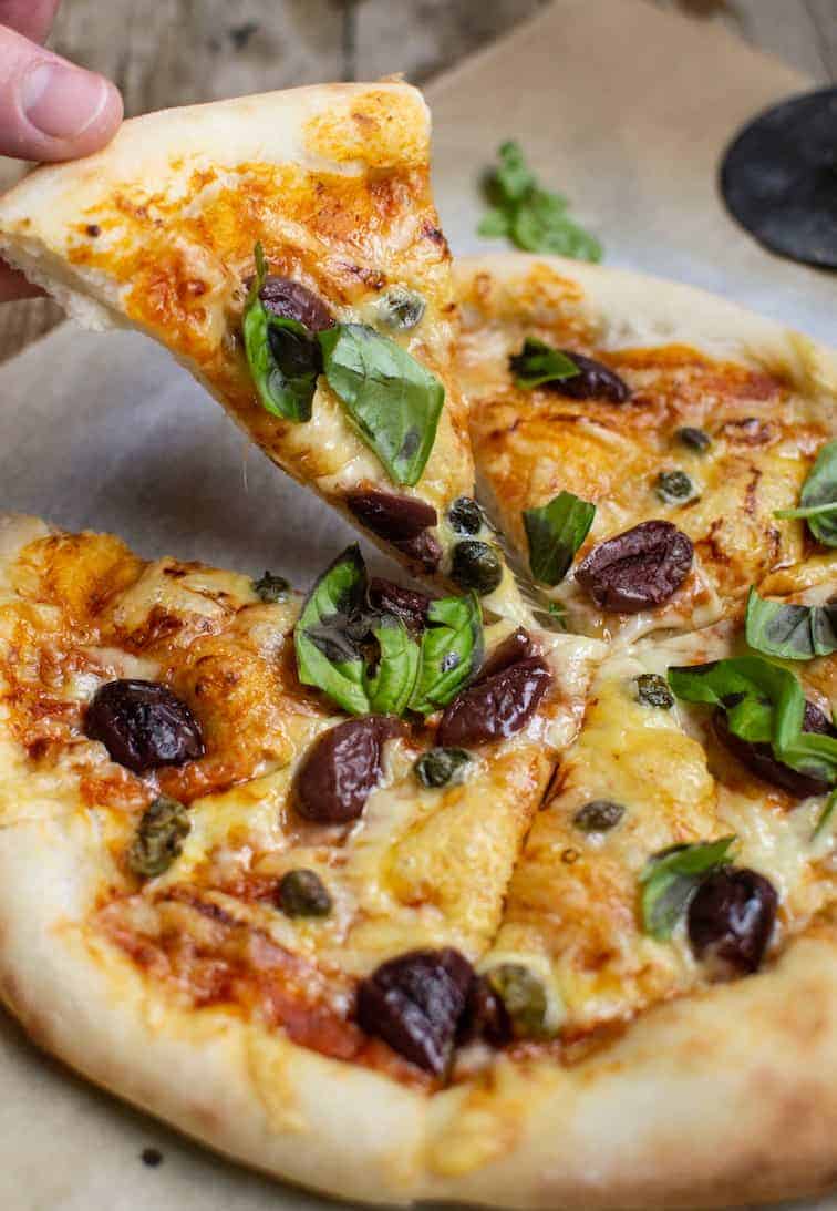 someone grabbing a slice of sourdough pizza with cheese, olives and basil topping