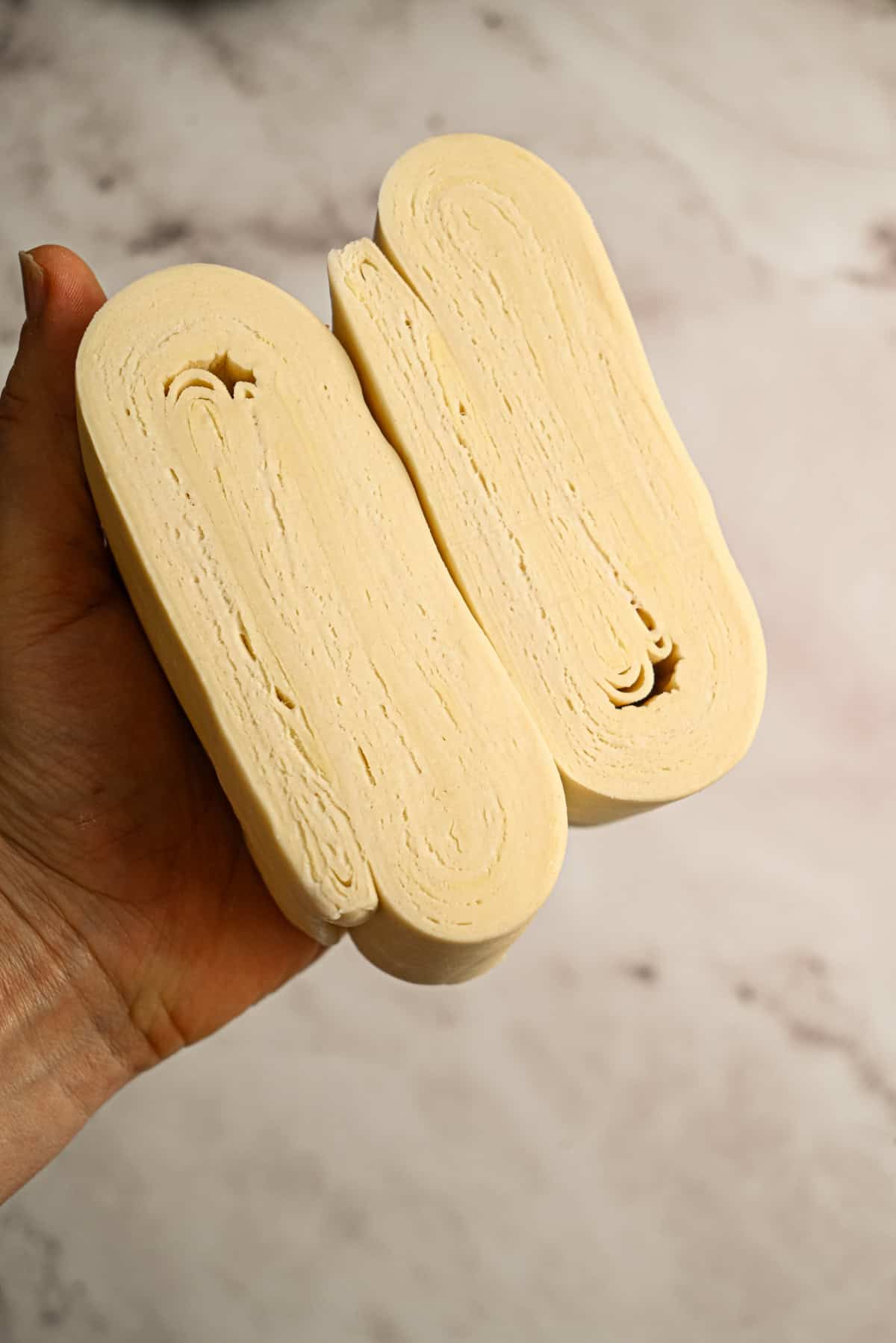 A hand holds a block of sourdough puff pastry cut in half, showing thin, even layers inside against a light, slightly marbled background.