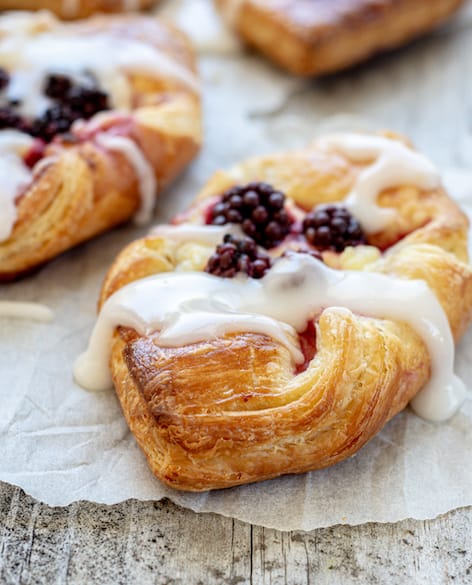 A close up of sourdough Danish pastries with 4 blackberries on it, on baking paper and drizzled with white icing