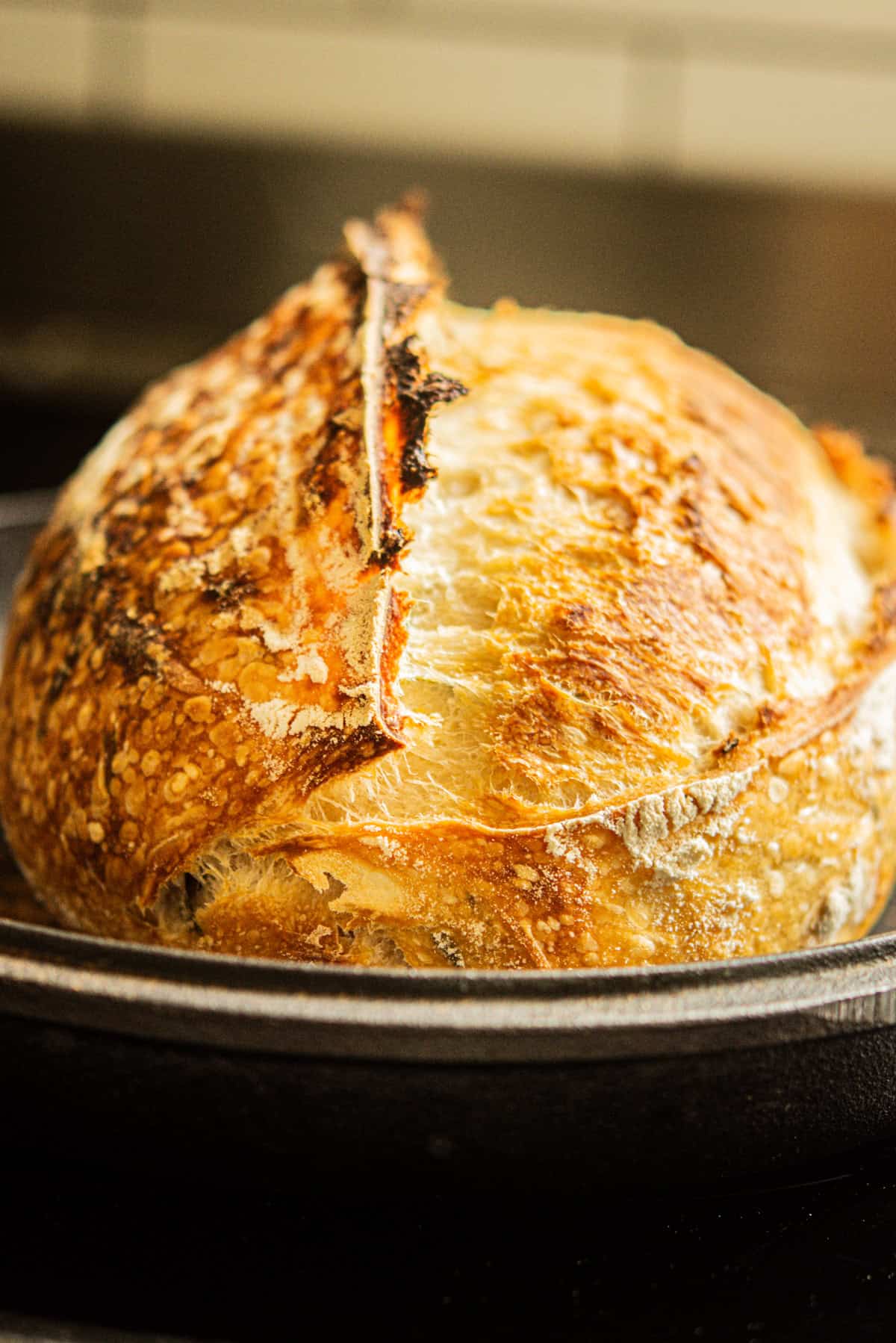 A round loaf of crusty, golden-brown sourdough bread sits in a cast iron skillet, its split top revealing a dense, textured surface in a warmly lit kitchen setting.