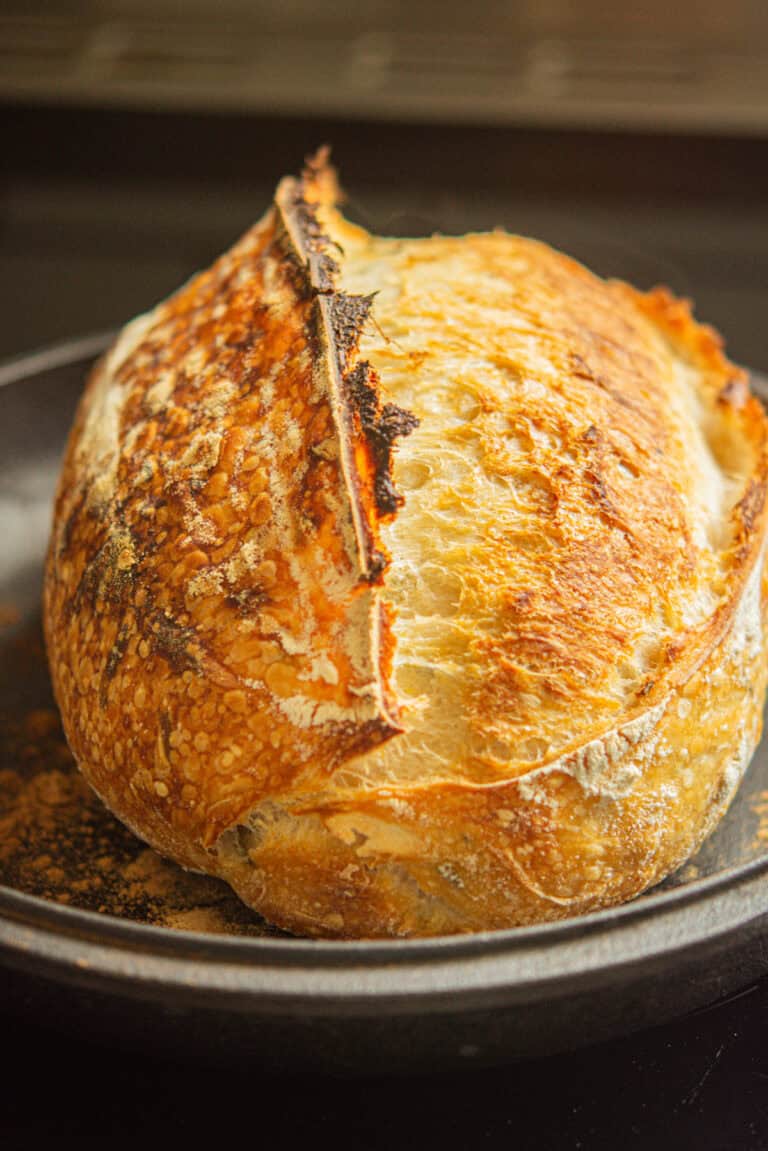 A round, golden-brown loaf of artisan sourdough bread with a crispy, crackly crust sits on a dark plate. The sourdough loaf has a dense interior, prominent score mark, and a rustic, homemade appearance.