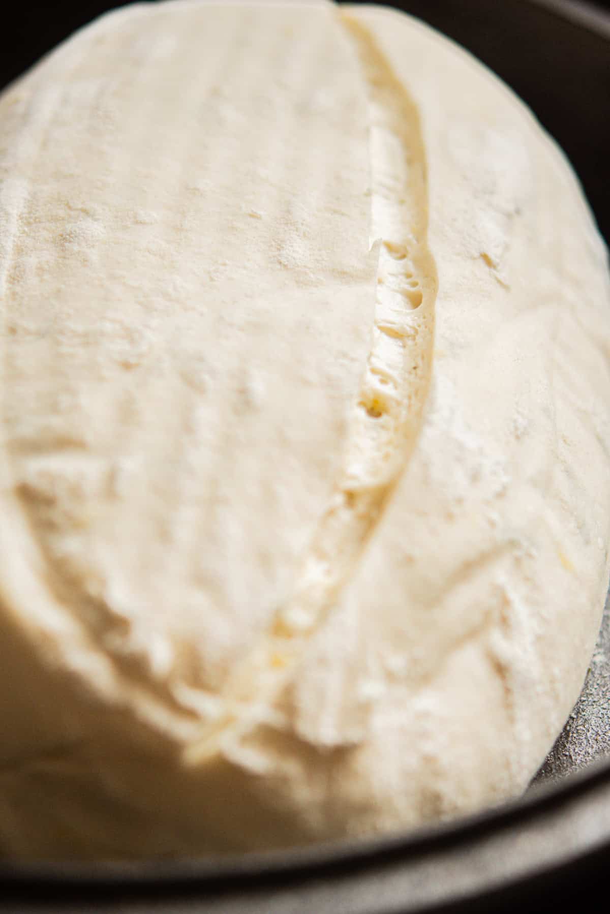 A close-up of a dense sourdough loaf of raw, scored bread dough in a pan, ready to be baked. The surface is dusted with flour and features a deep slash down the middle.
