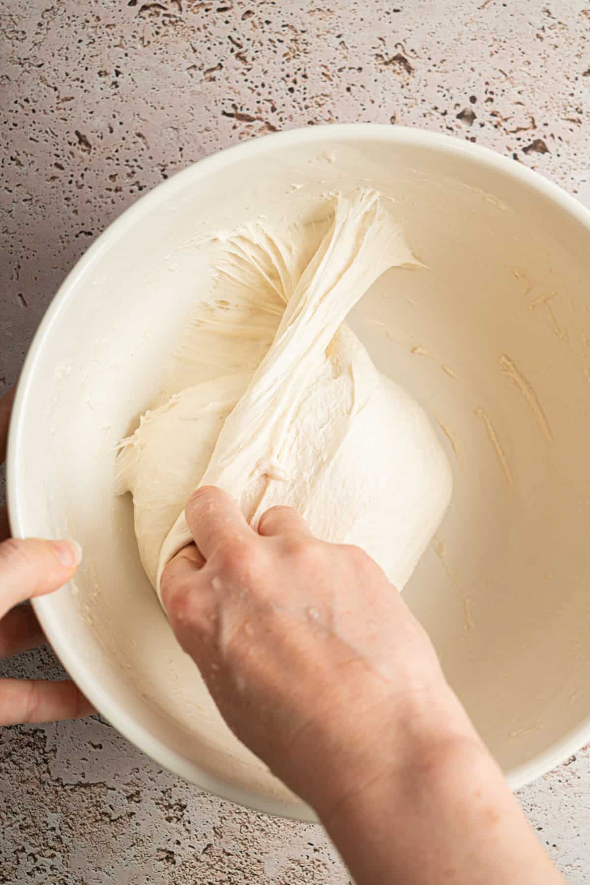 A person’s hands stretch and fold dense sourdough bread dough inside a large white mixing bowl on a speckled countertop.