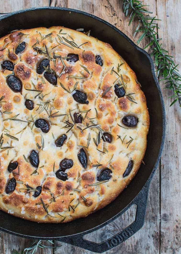 a cast iron pan with baked focaccia bread in it with olives and rosemary 