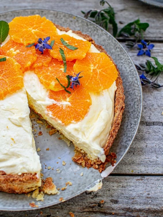 lemon yogurt tart with sliced oranges and mint on top, with one slice missing on a grey plate and on top of a wooden table