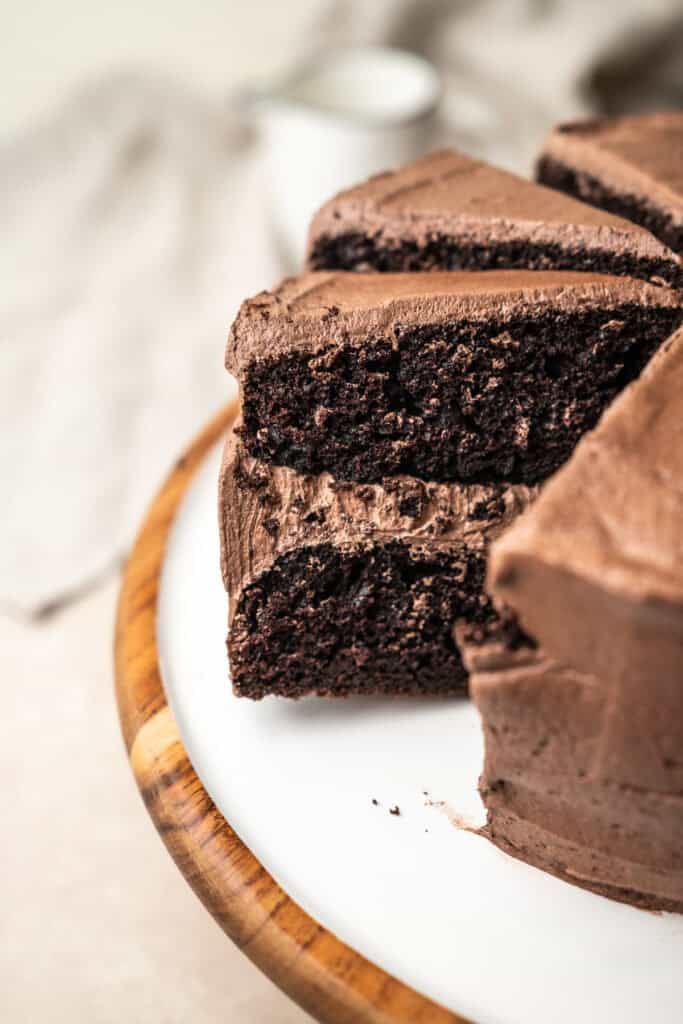 A close-up of a sliced sourdough chocolate cake with rich chocolate frosting on a white plate, sitting on a wooden cake stand.