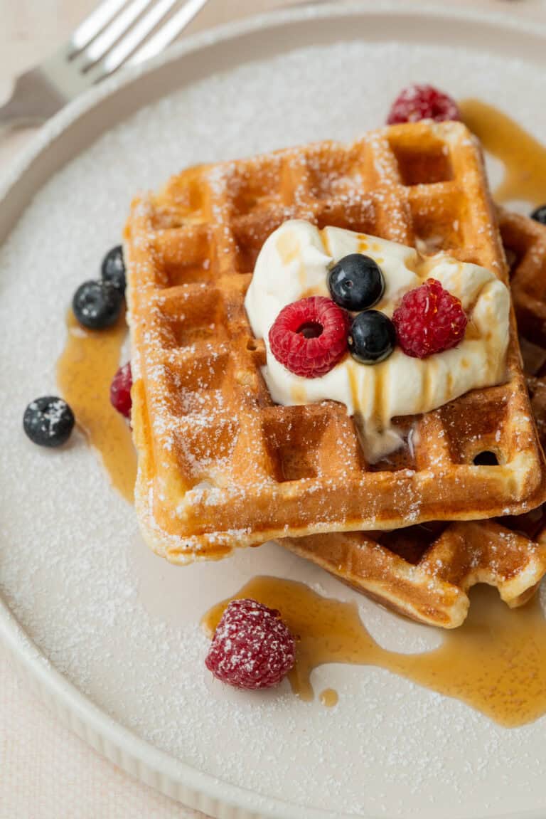 Two golden Sourdough Discard Waffles topped with whipped cream, blueberries, raspberries, and a drizzle of syrup sit on a white plate dusted with powdered sugar. More berries are scattered around, and a fork is visible in the background.