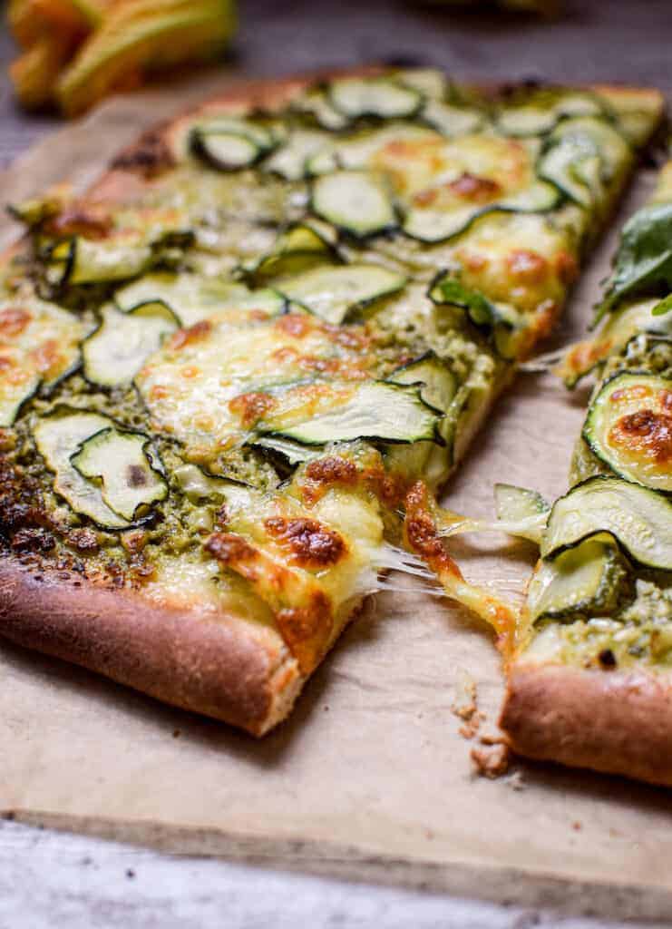 a side view of a zucchini pesto pizza, a piece being picked up with melted cheese stretching off it 