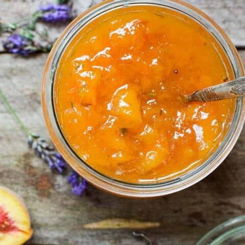 Birds eye view of a jar filled with orange peach preserve with thyme leaves and a spoon handle poking out the top of the jar. The jar is on top of a wooden background with lavender flowers scattered around a half peach and a jar lid.