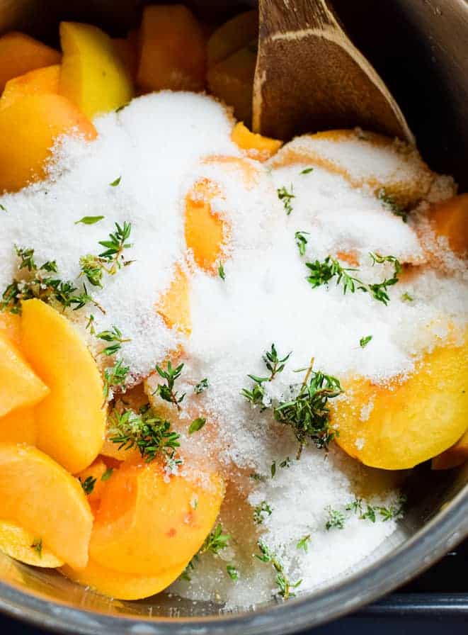 Chunks of peach in a glass bowl half covered with sugar and thyme. A large wooden spoon in the bowl at the top