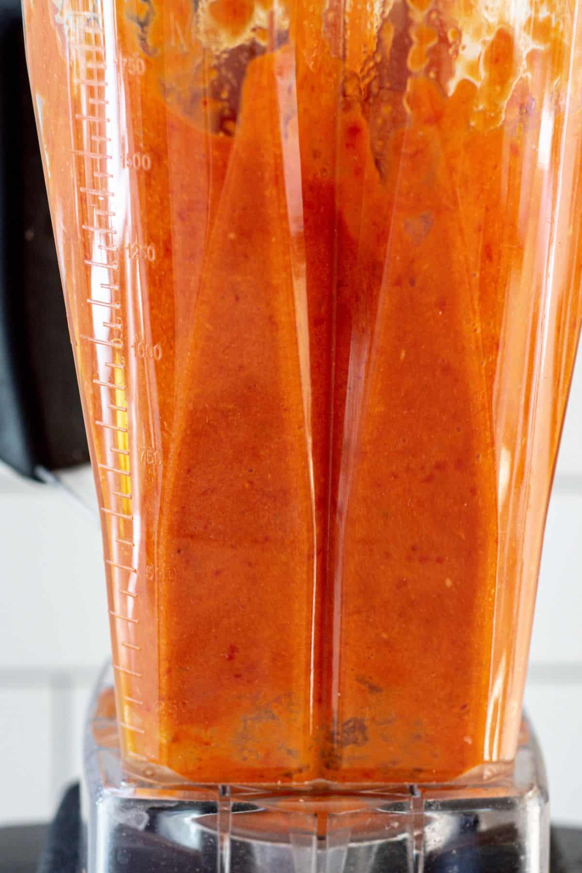 A close-up view of a blender filled with smooth, bright orange-red fermented hot sauce being blended, with measurement markings visible on the side of the container.